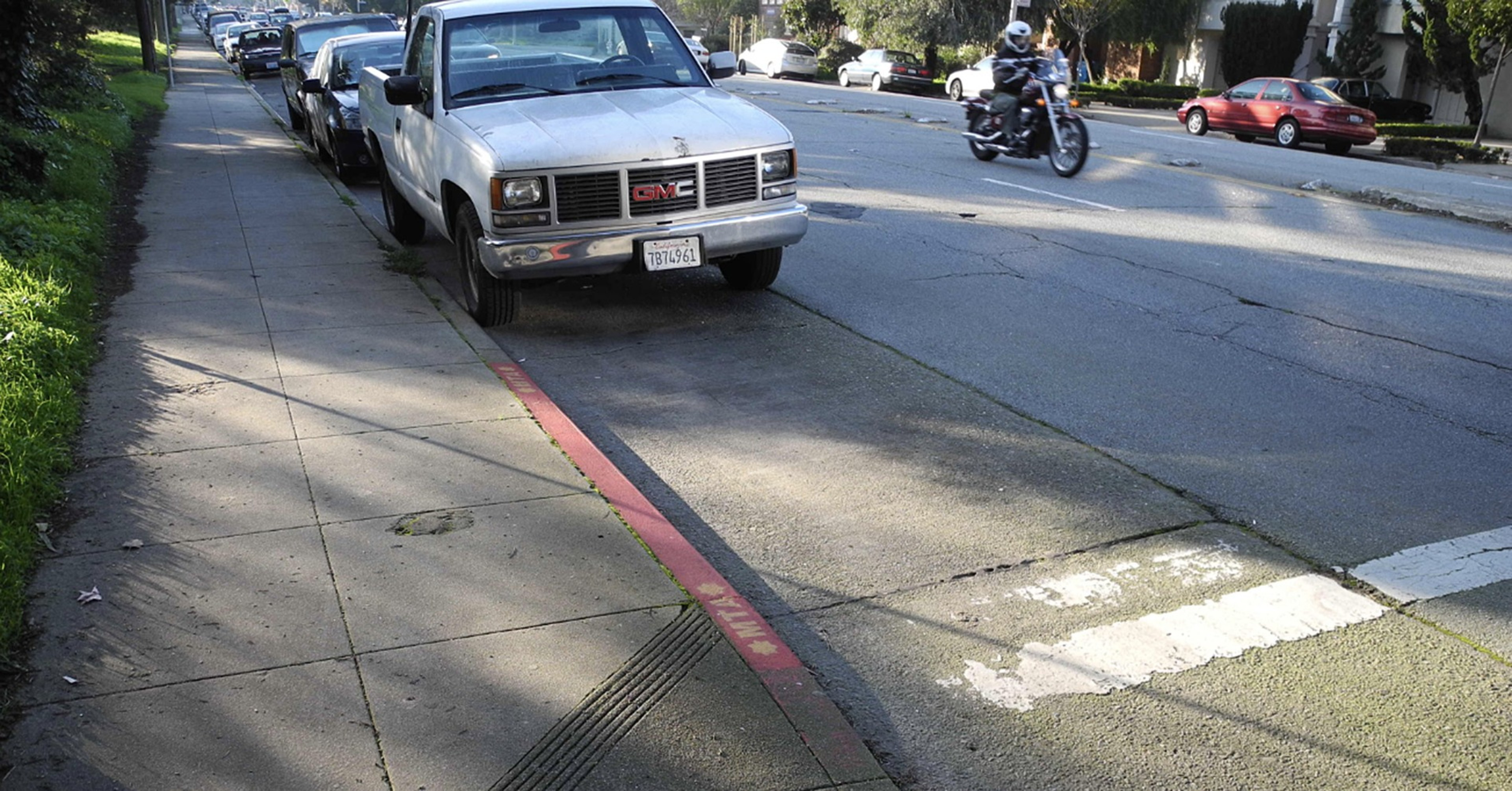 A truck is parked on a sidewalk next to a street where a motorcycle passes by. There's a red curb and greenery along the walkway.