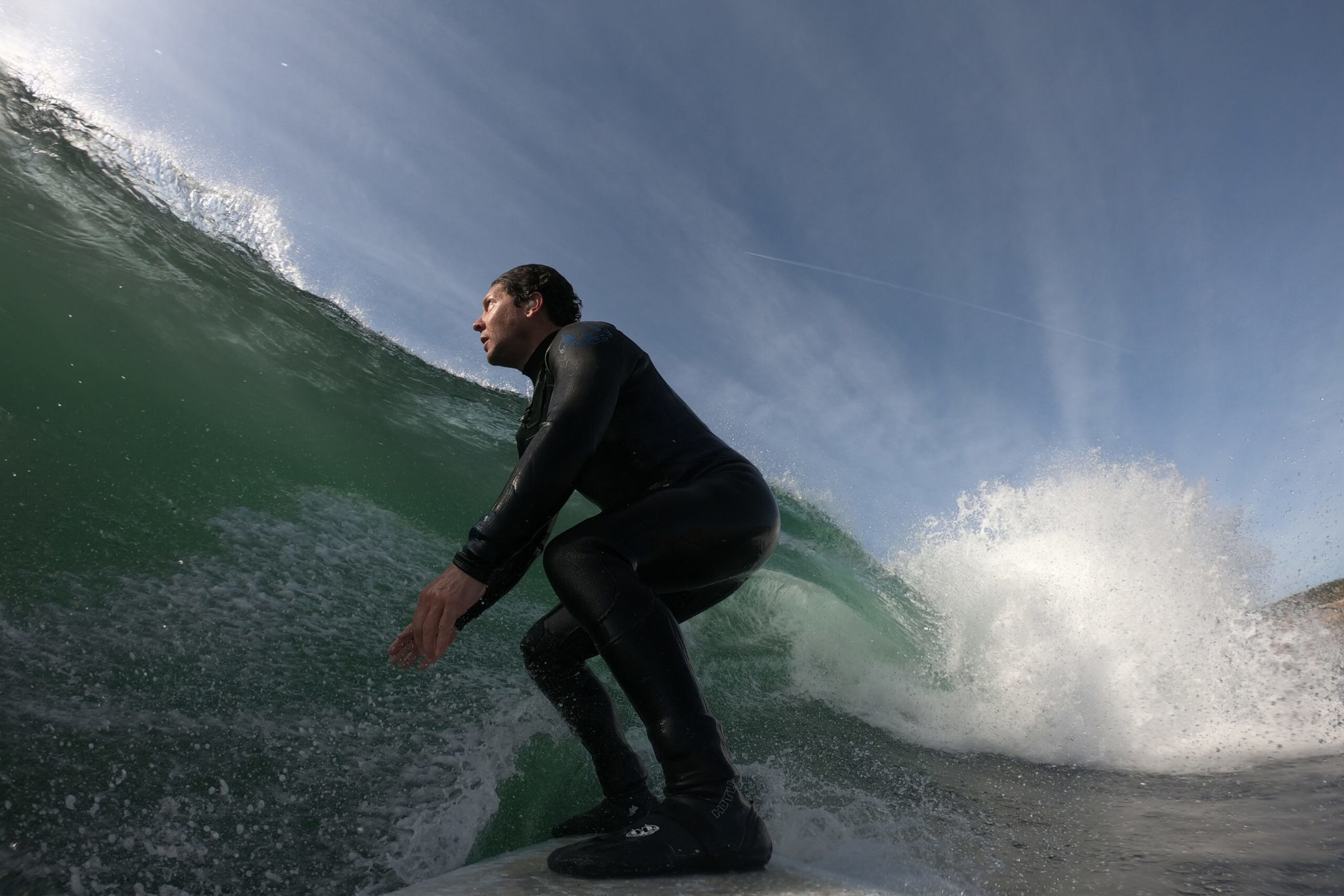 A surfer in a black wetsuit rides a large, curling green wave under a clear blue sky.