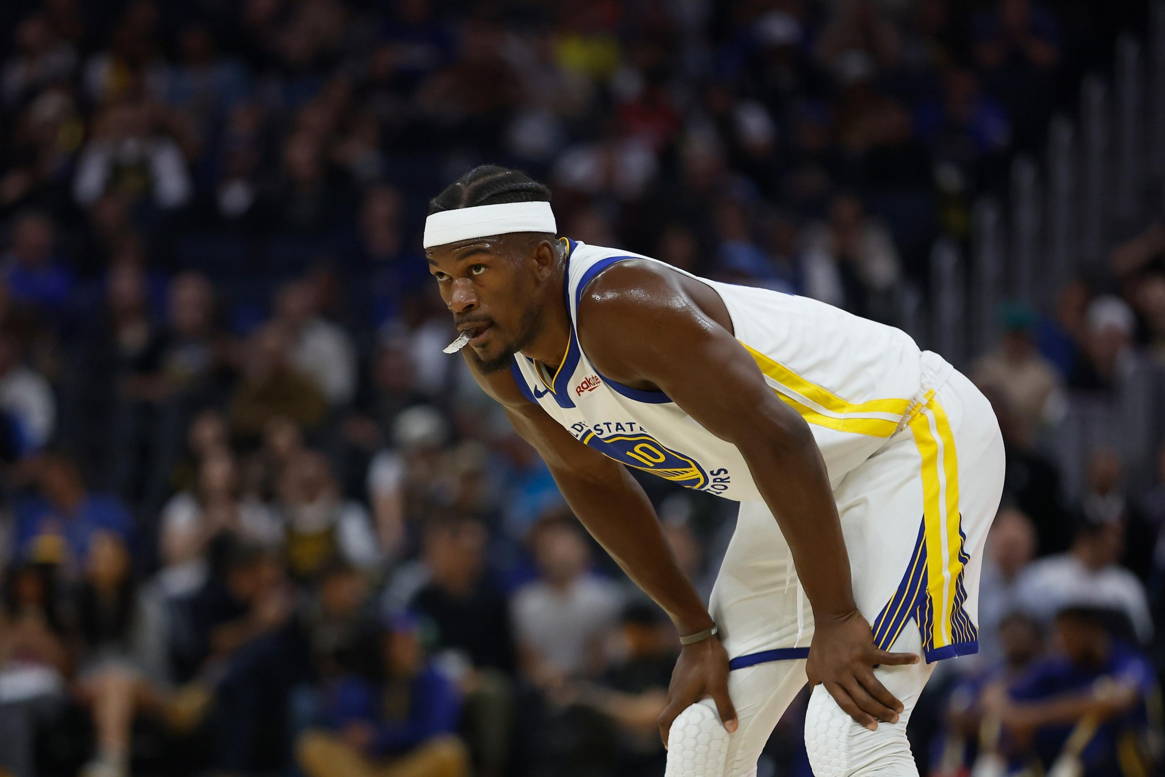 A basketball player in a white Golden State Warriors jersey with number 10 leans forward, resting his hands on his knees during a game.
