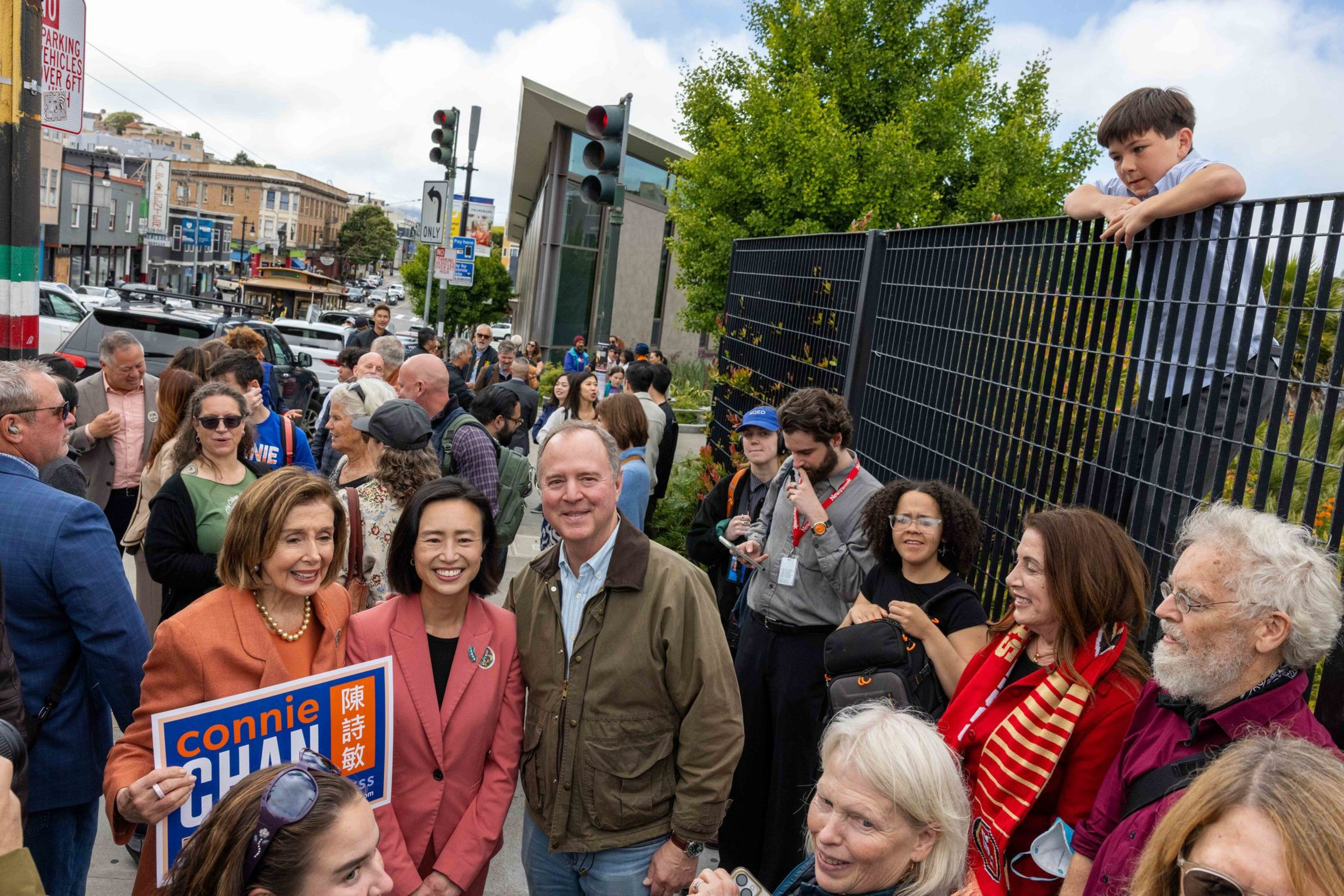 A diverse group of people gathers outdoors near a street, with some smiling at the camera; a boy leans on a black fence above the crowd.