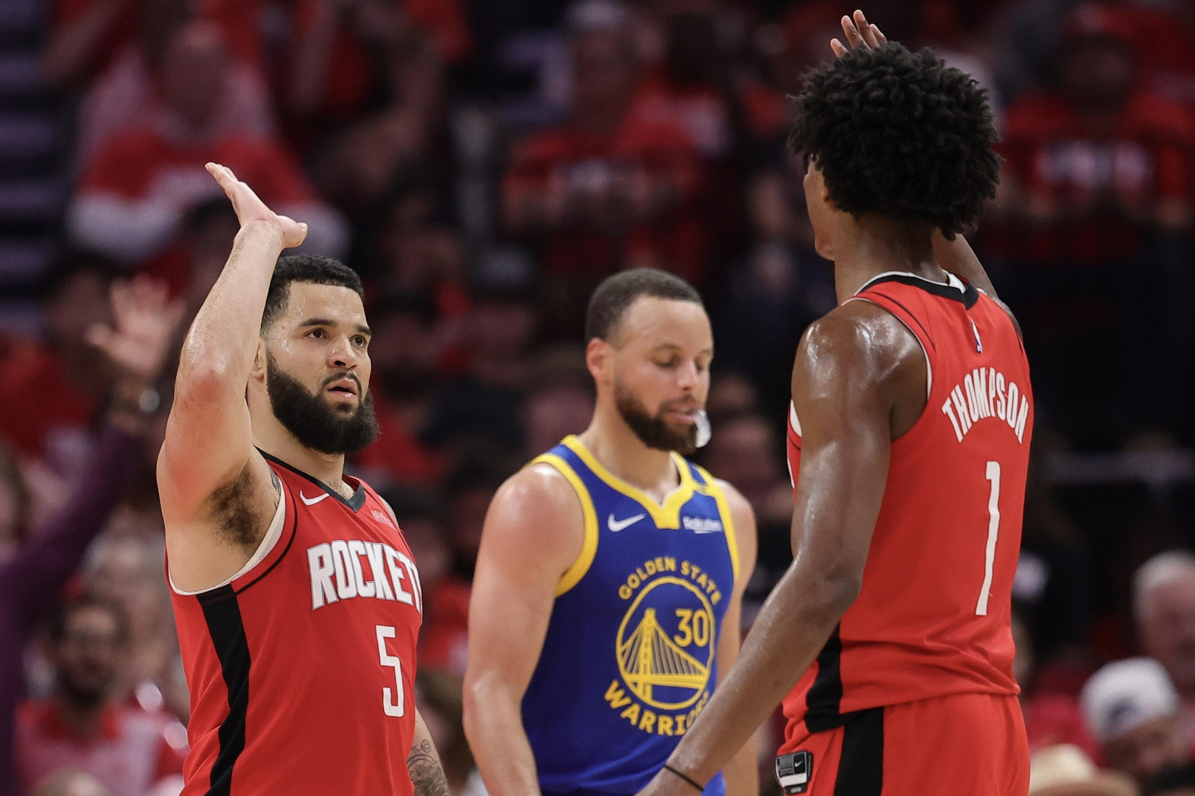 Two basketball players in red jerseys high-five, while a player in a blue jersey walks past them. The background shows a cheering crowd.