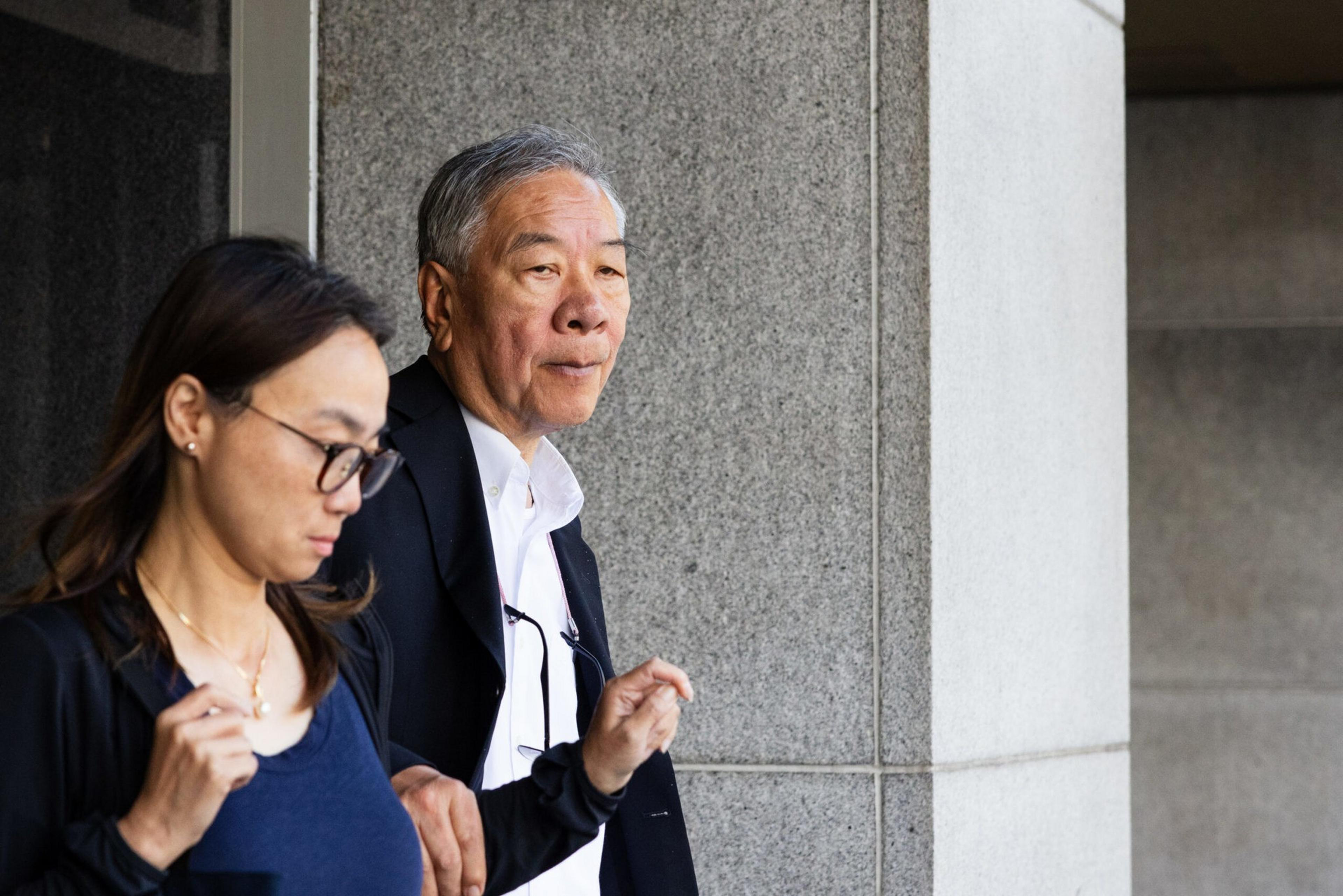 An older man and a younger woman, both wearing dark jackets, stand near a stone wall, looking in different directions with serious expressions.