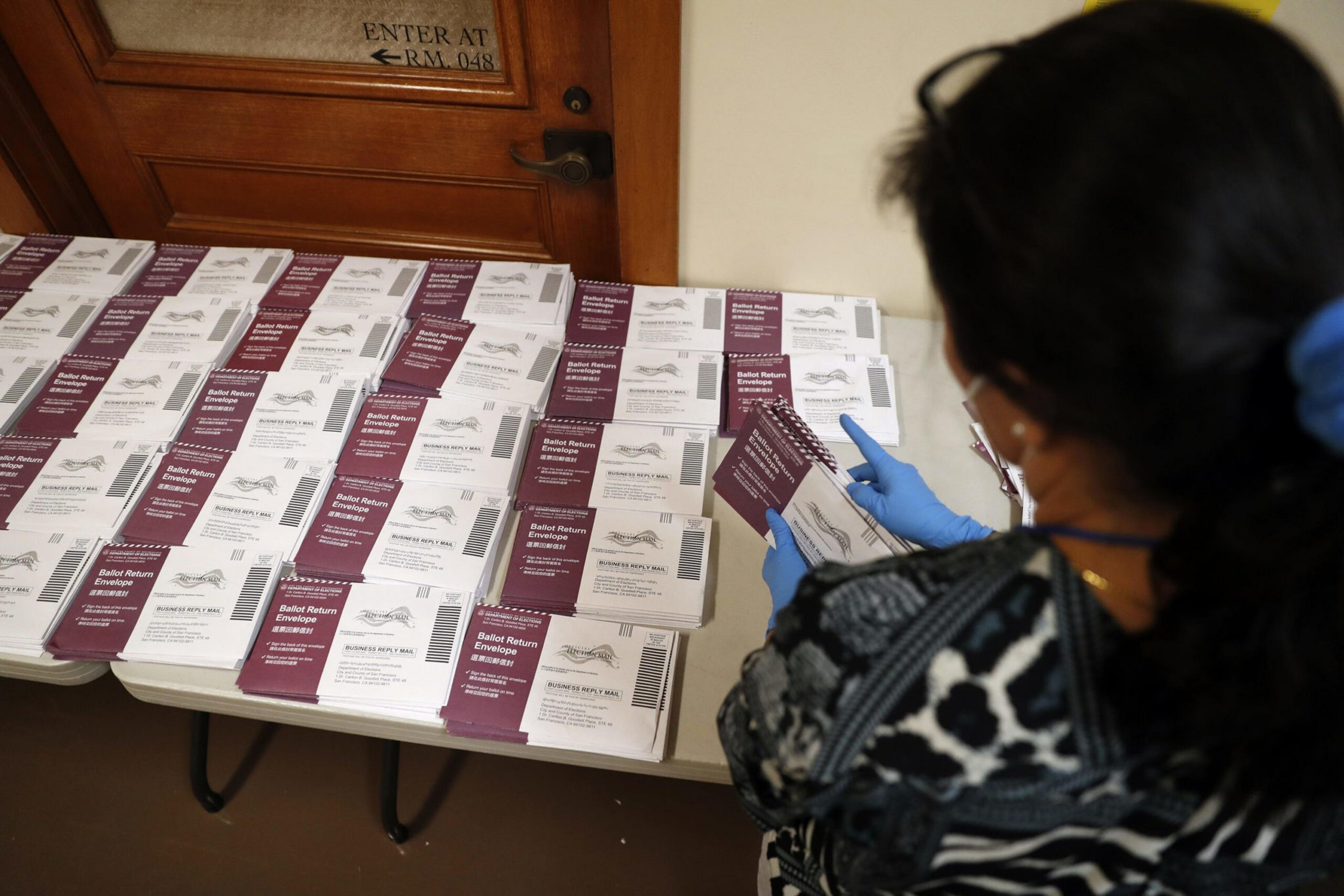 A person wearing blue gloves sorts stacks of maroon and white ballot return envelopes laid out on tables near a wooden door.