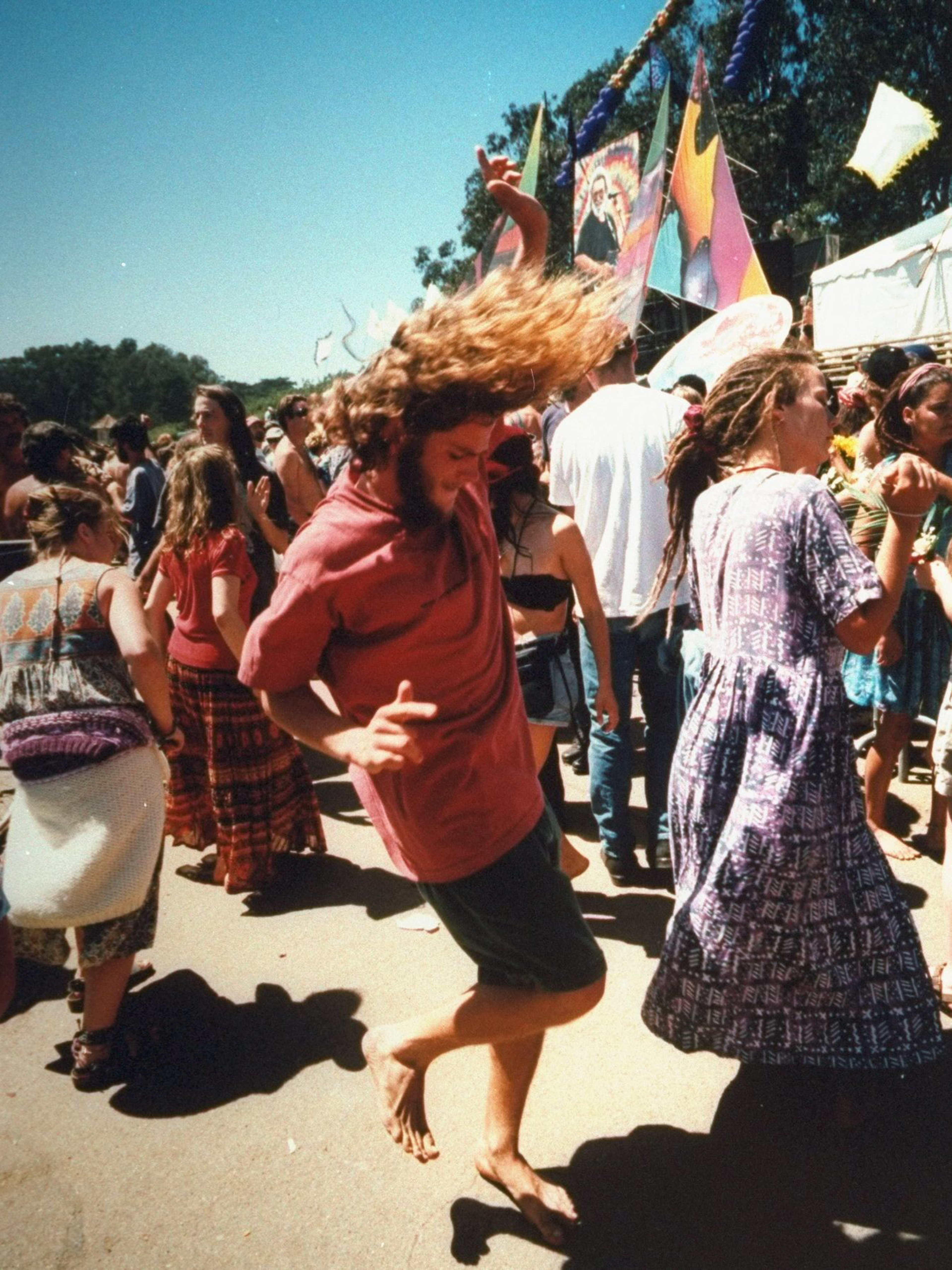 The image shows people dancing outdoors at a festival, with colorful banners and trees in the background. A man with long hair joyfully moves barefoot.