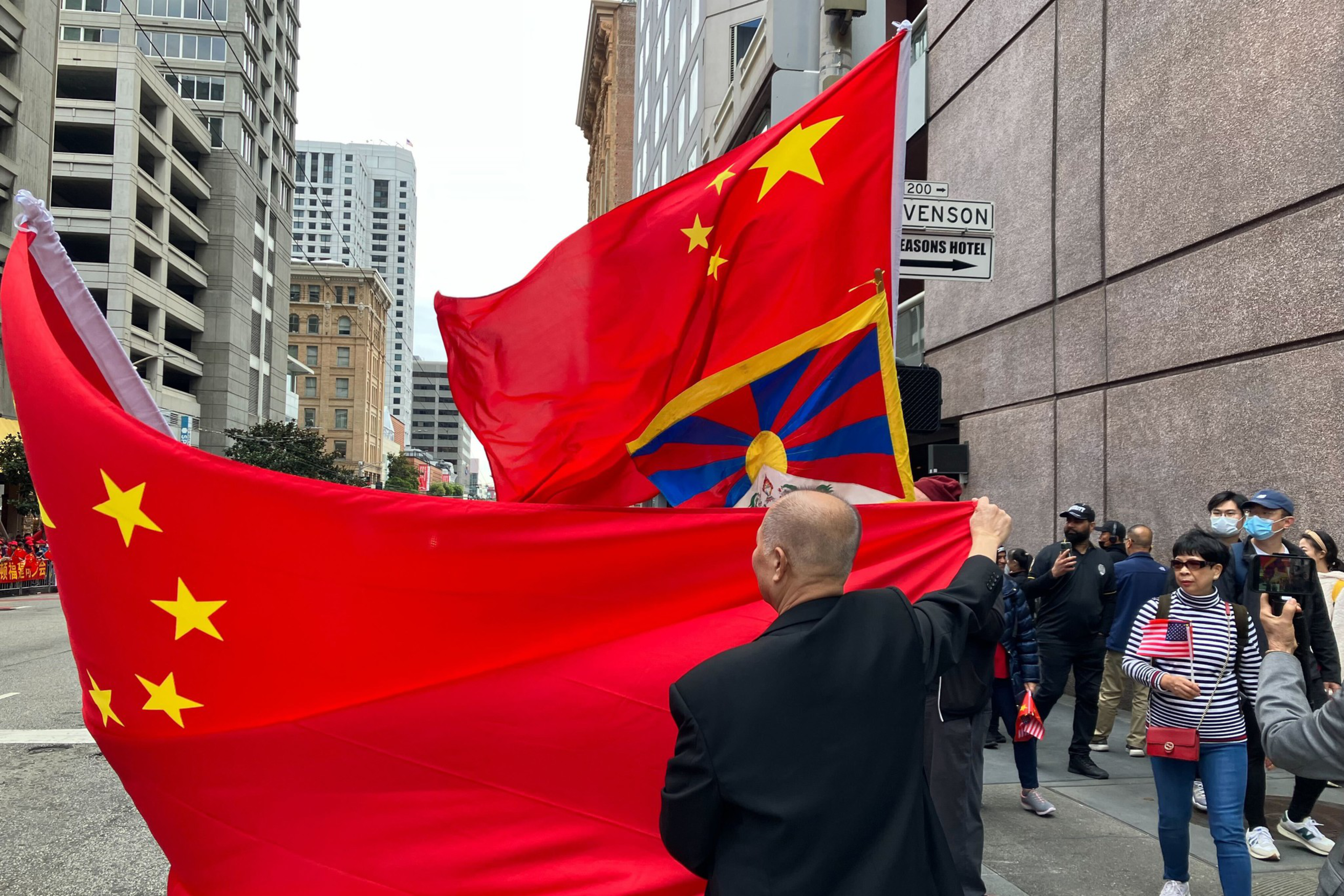 A man stretches a large, red, Chinese flag around a Tibetan flag on a city sidewalk as pedestrian pass.