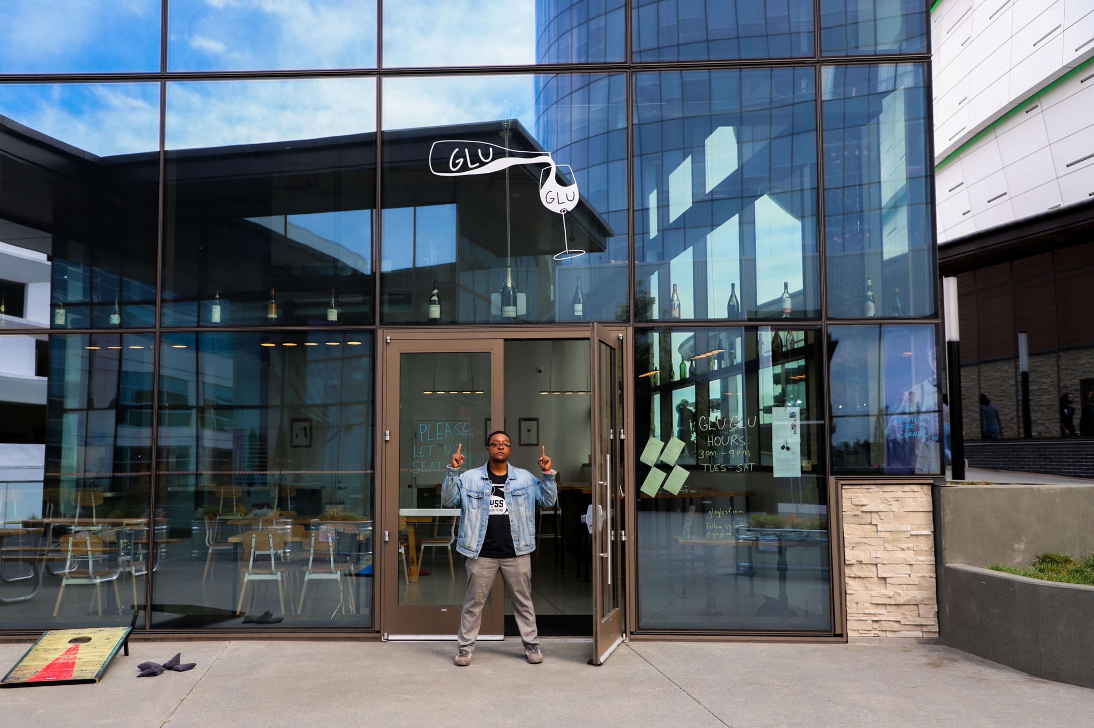 A person stands outside a glass-walled building with "GLU GLU" written above the entrance, wearing a denim jacket and gesturing with both hands.