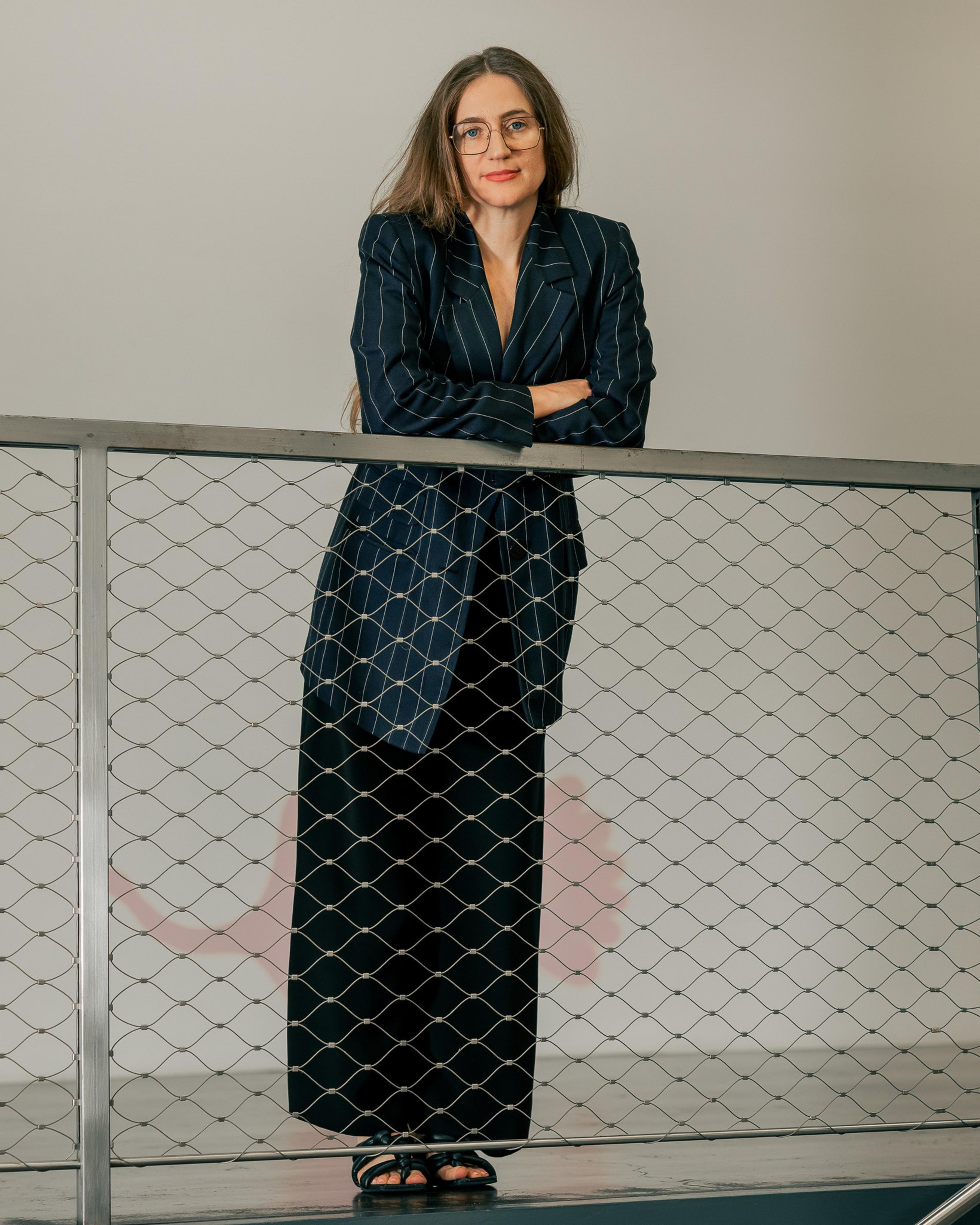 A person with long hair and glasses leans against a wire railing. They're wearing a pinstripe blazer and black pants, and the background is plain.