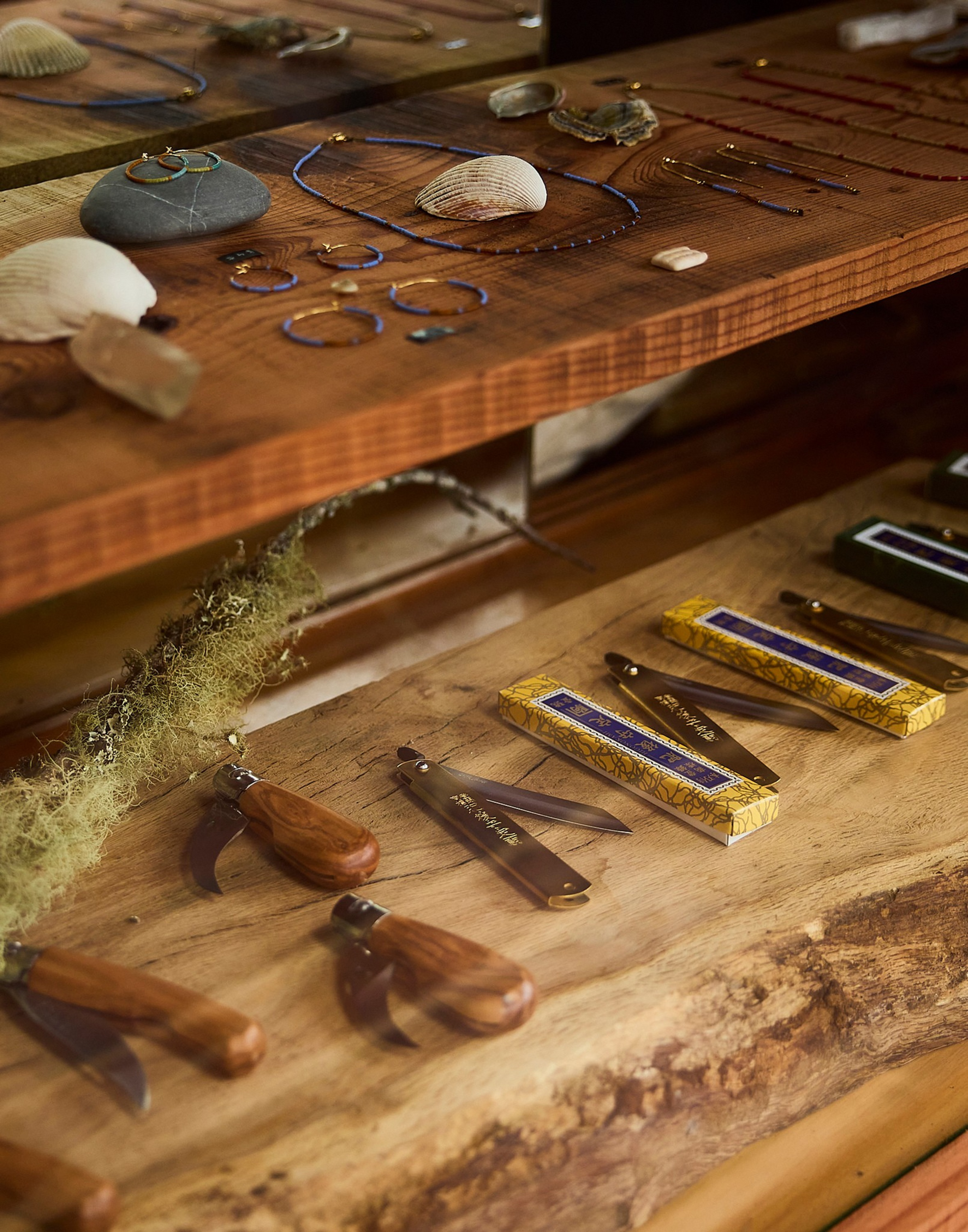 Wooden-handled folding knives and decorative keychain knives are arranged on a wooden surface, with jewelry and seashells on a shelf above.
