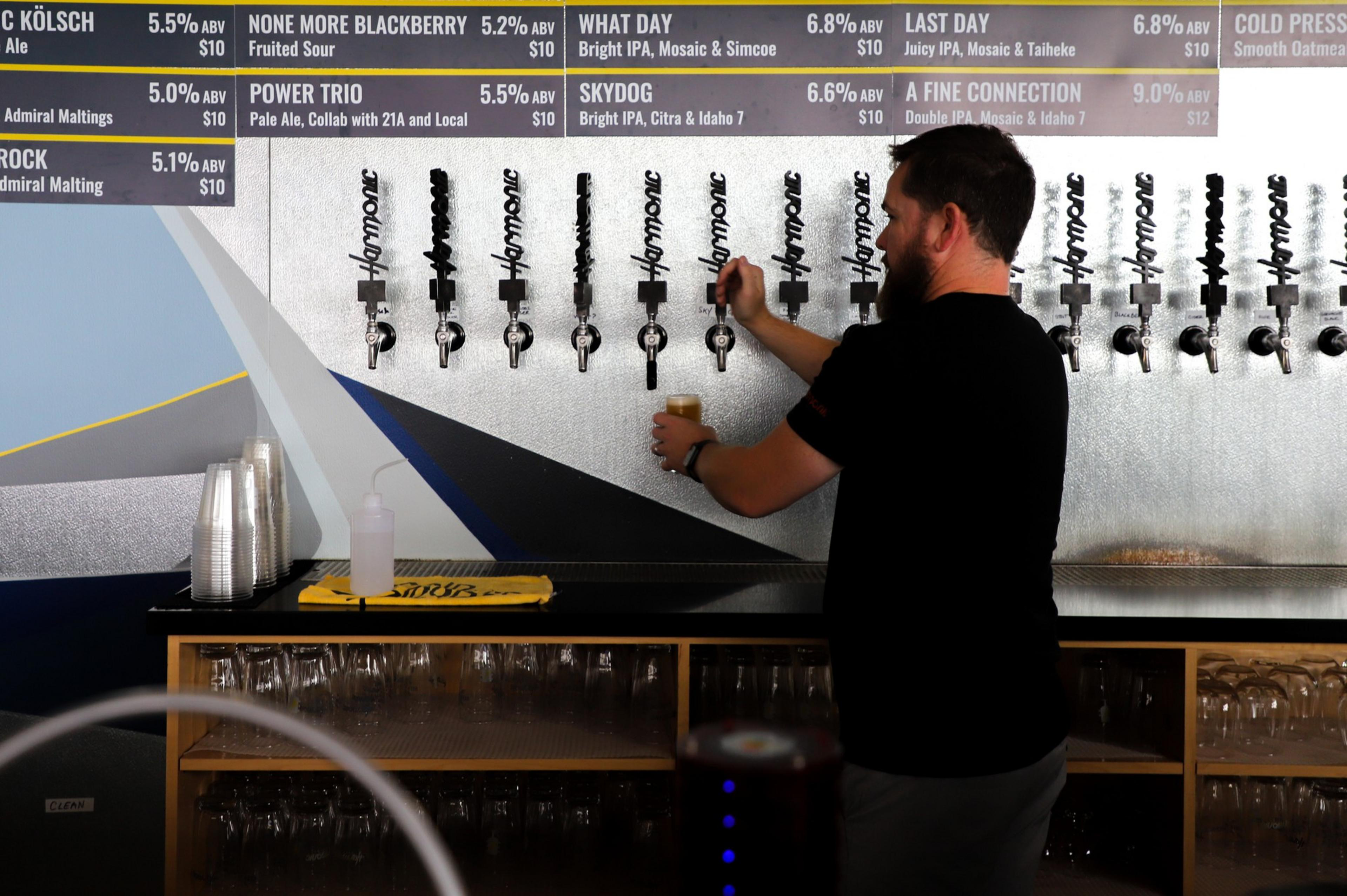 A man with a beard fills a glass from one of several beer taps mounted on a metal wall beneath a menu listing various beer names, ABVs, and prices.