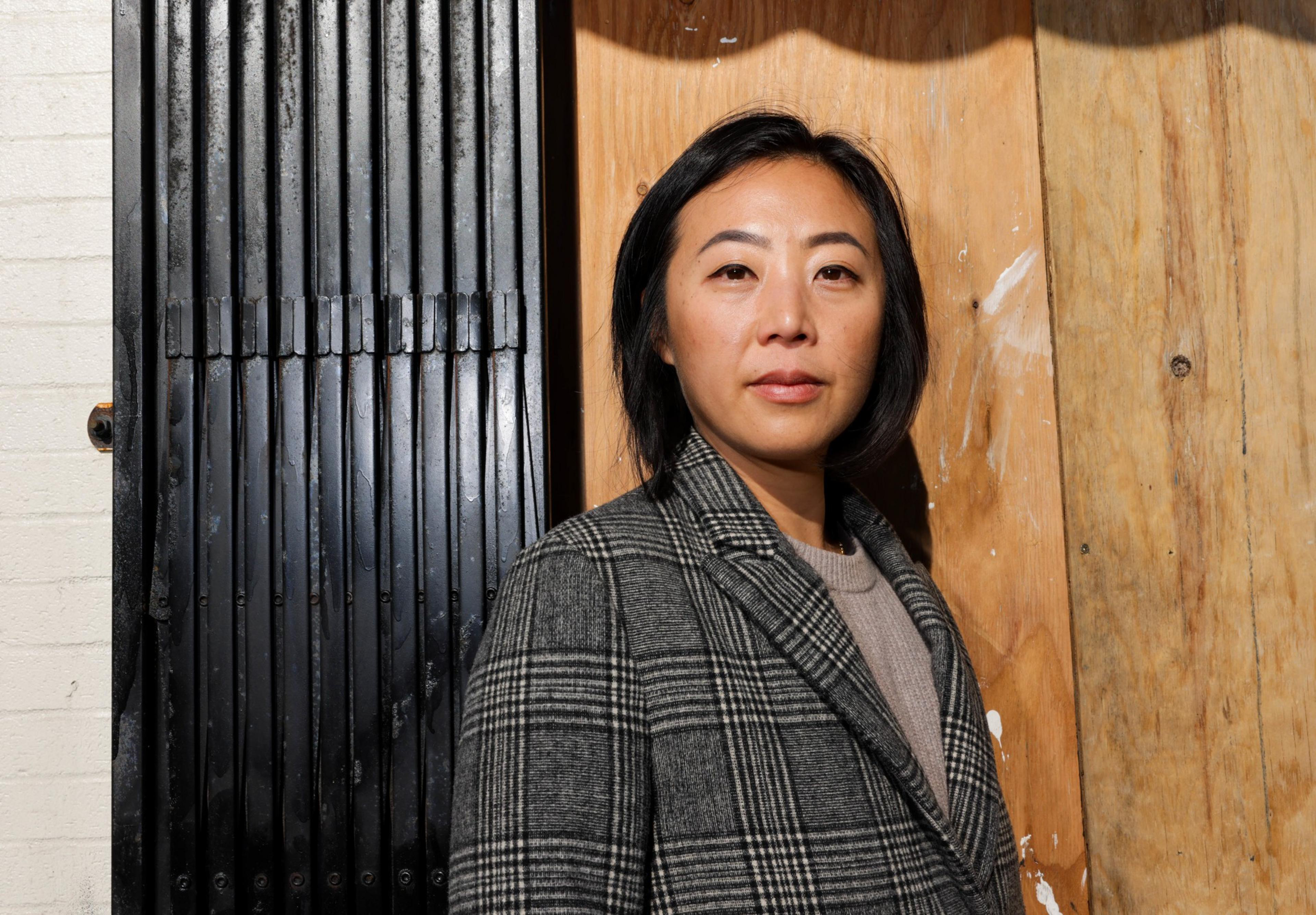 A person in a gray overcoat stands in front of their business that is boarded up with plywood.