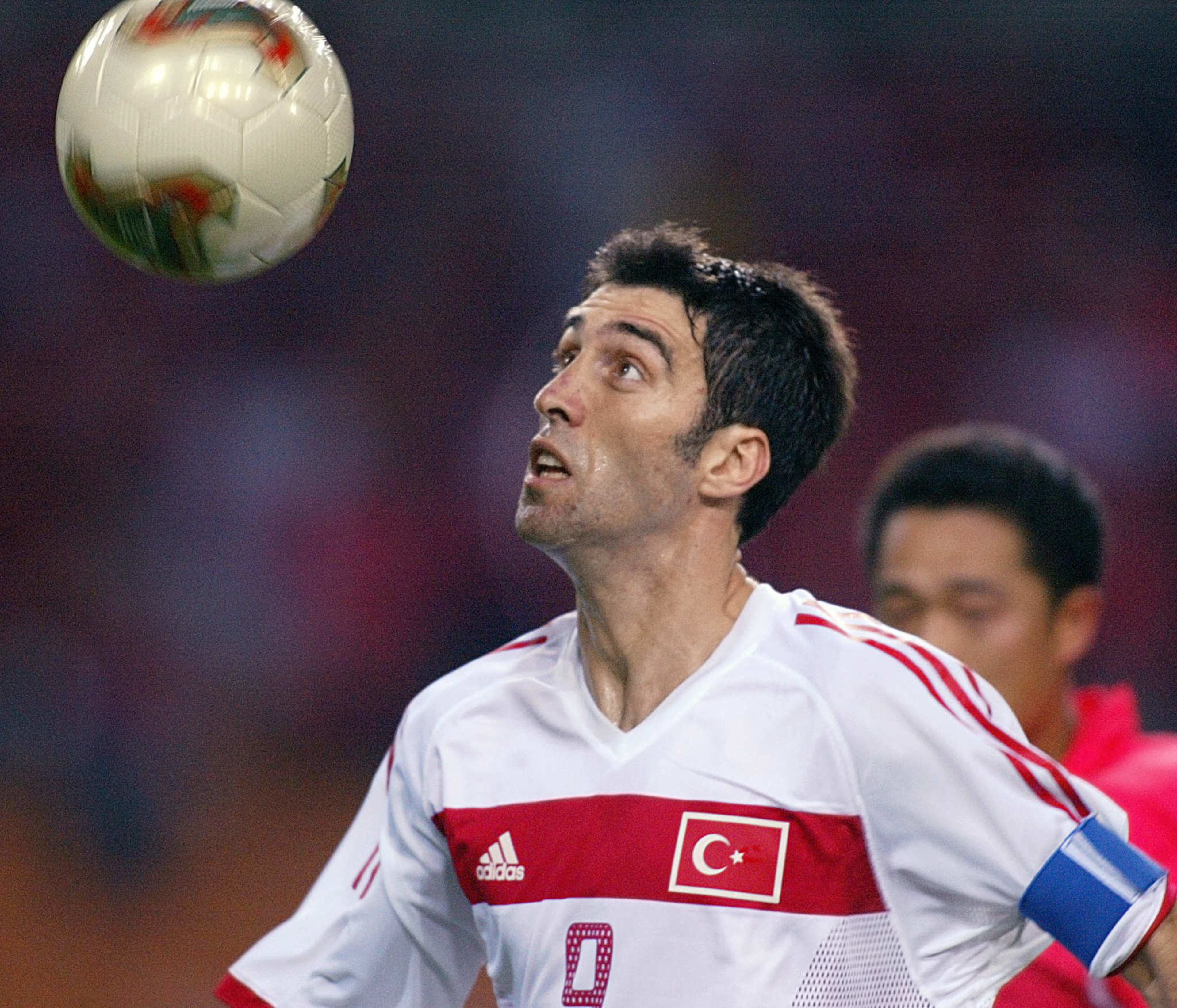 A soccer player wearing a white jersey with Turkey’s flag and captain’s armband looks up at a ball in midair during a match.