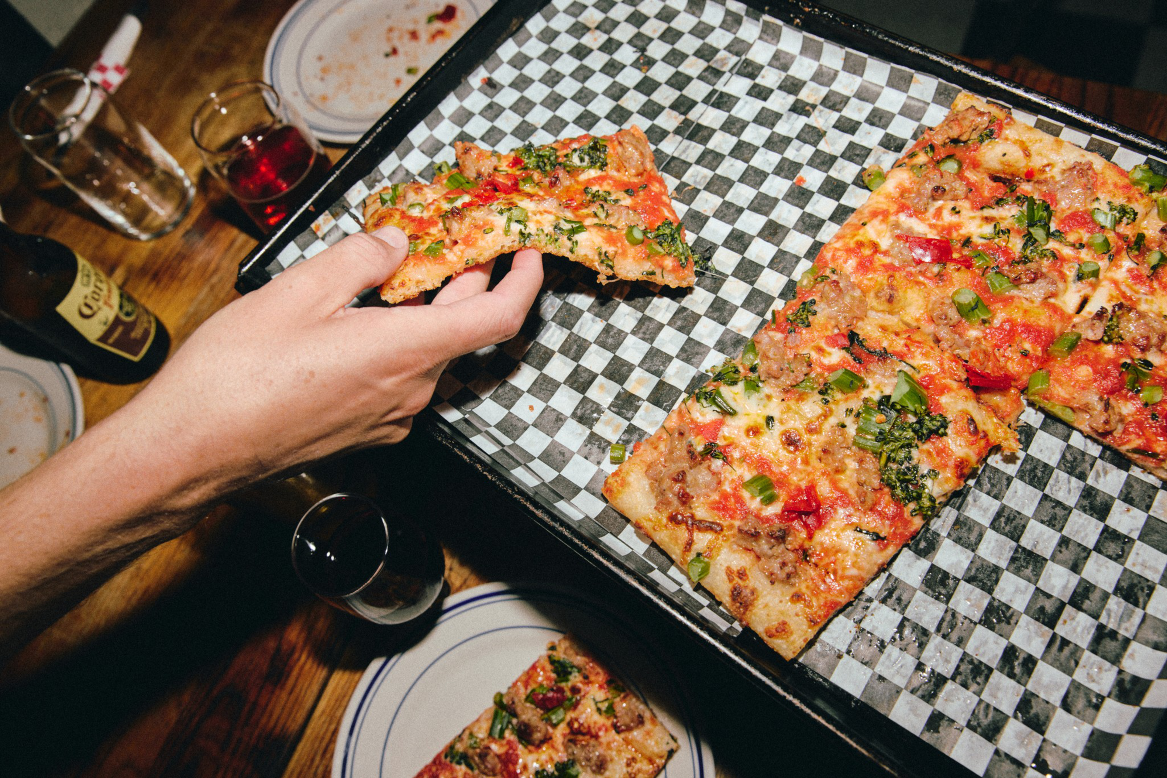 A hand is picking up a slice of rectangular pizza with vegetables and sausage from a checkered paper-lined tray, with drinks and plates on a wooden table.