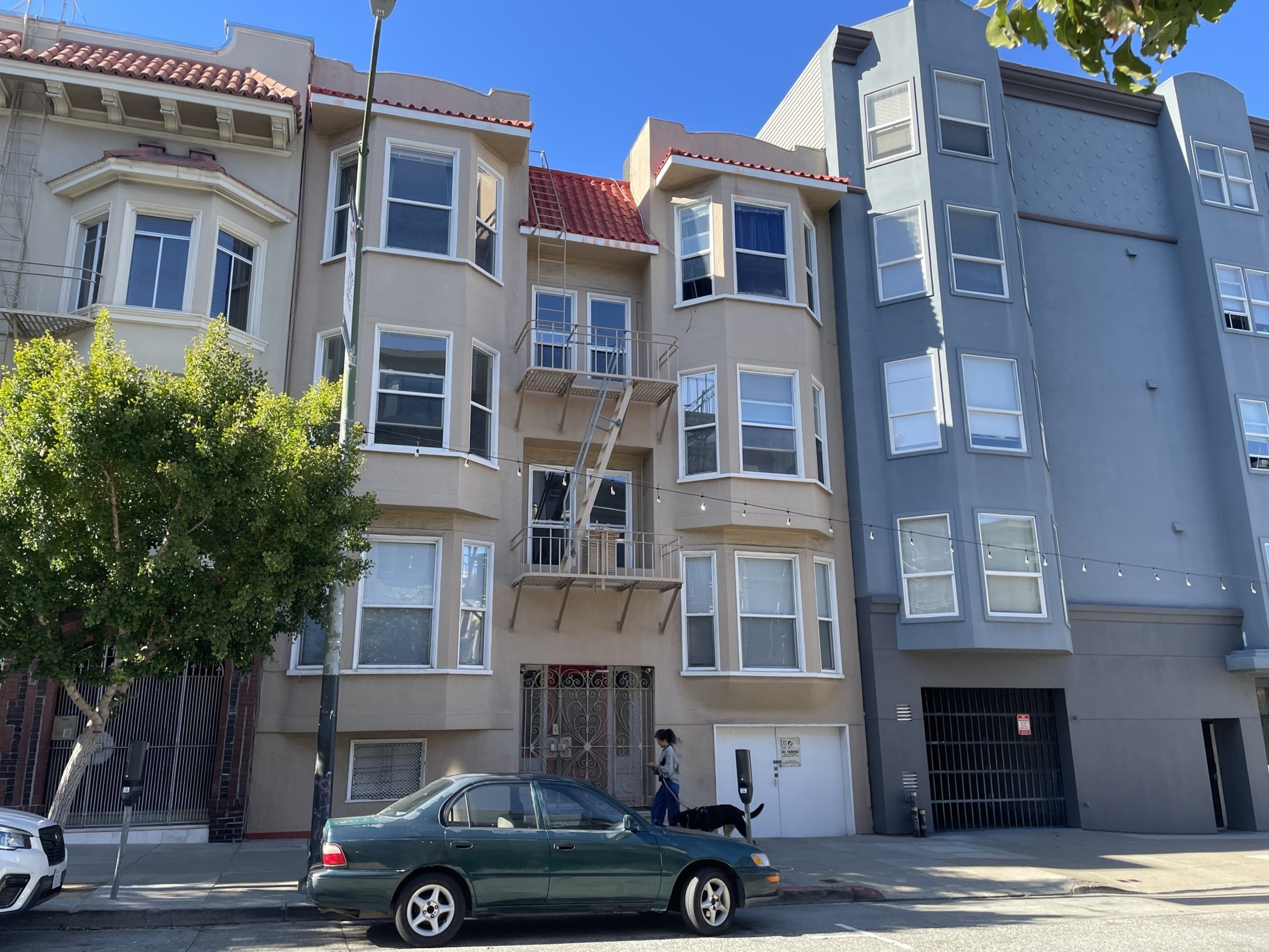 Three-story beige and gray buildings with bay windows, a small tree near the curb, a green sedan parked, and a person walking a black dog.