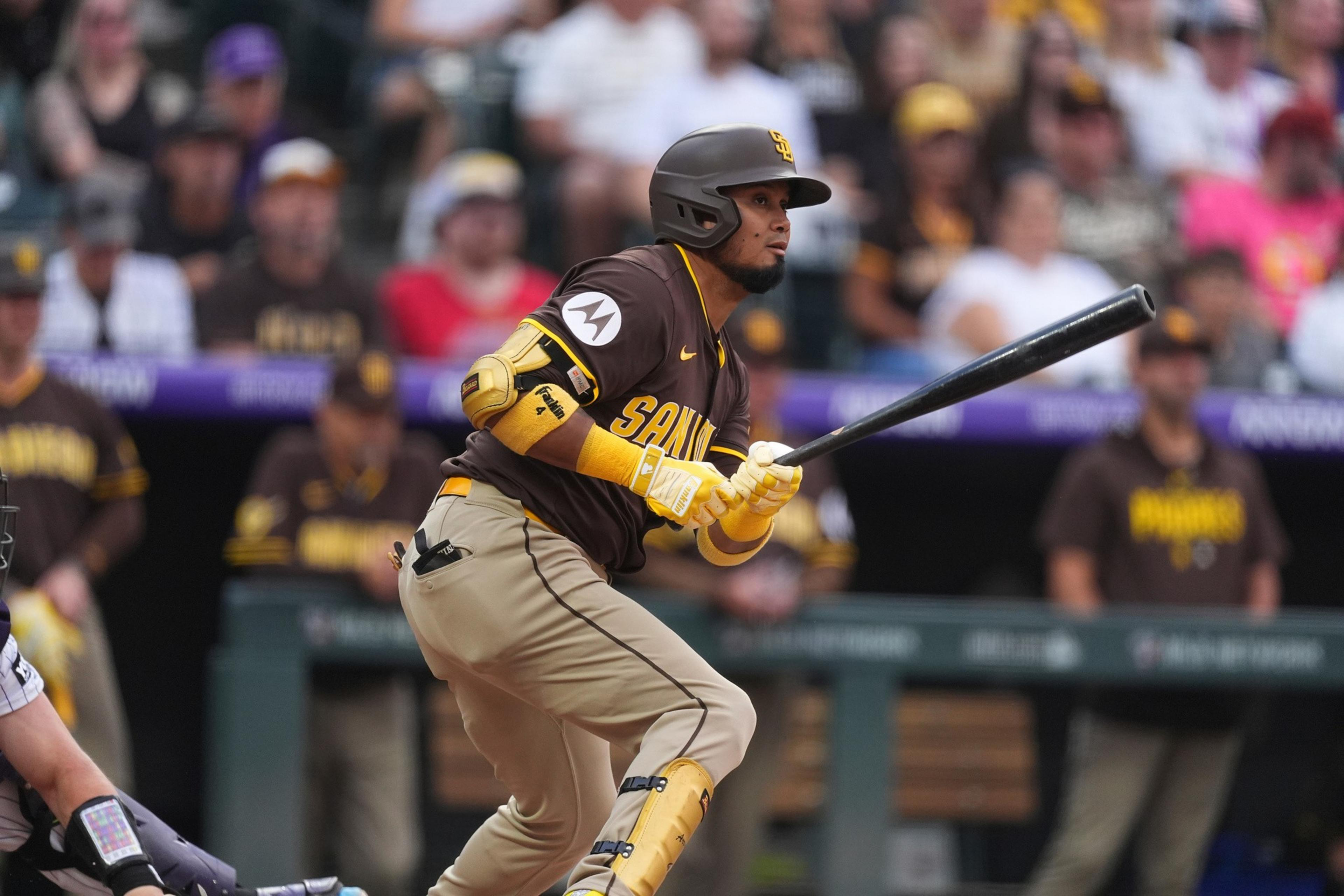 A baseball player in a brown and yellow San Diego Padres uniform swings a bat during a game, with fans and teammates blurred in the background.