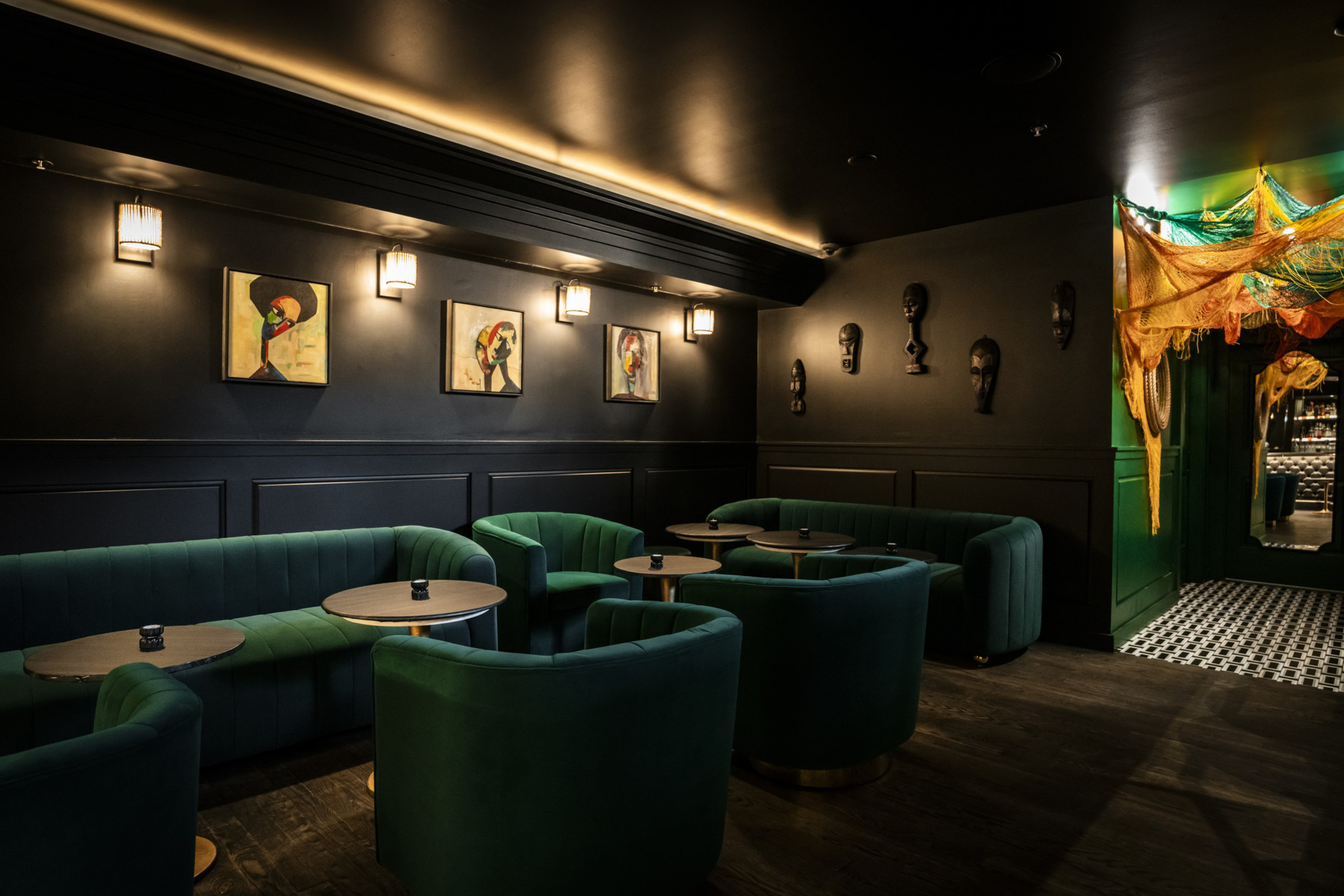 The image shows a dimly lit lounge with green velvet seating, small round tables, and modern artwork on dark walls. Masks and colorful netting adorn the space.