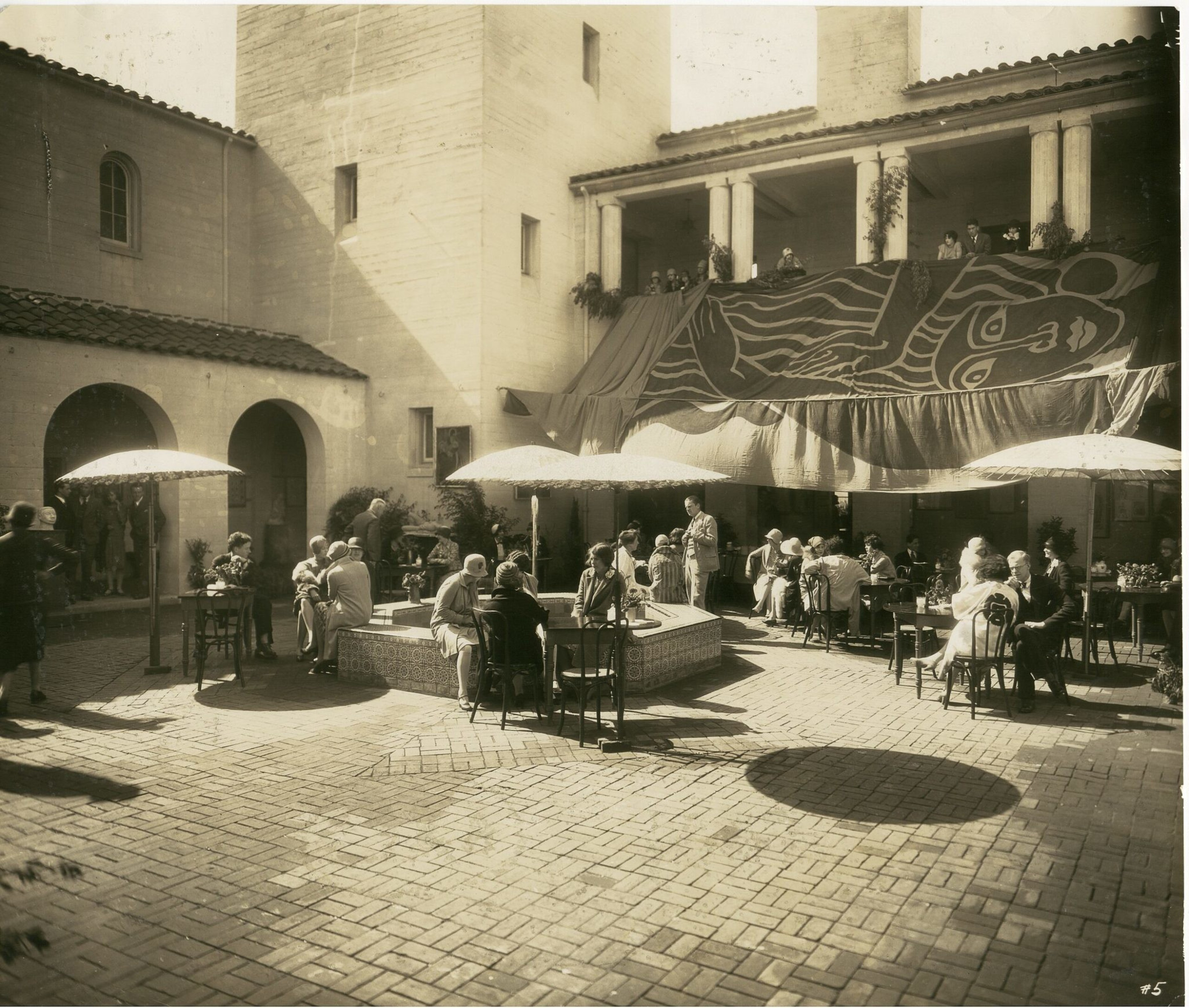 People sit and chat at tables with umbrellas in a sunlit courtyard featuring a central fountain and a large mural hanging from the second-floor balcony.