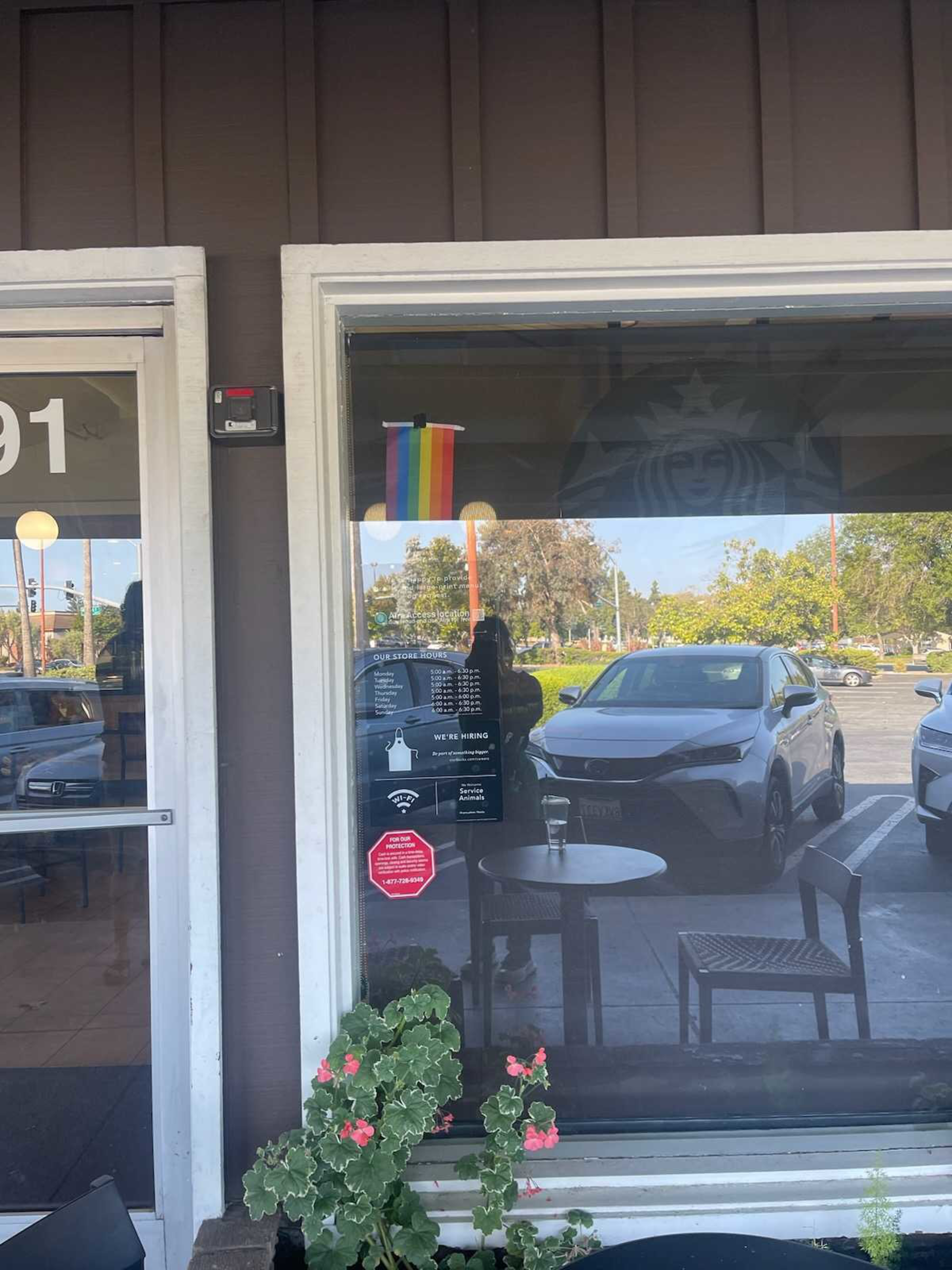A window displays store hours, hiring info, and a small pride flag, with a round table and chairs outside, reflecting cars and trees in the parking lot.