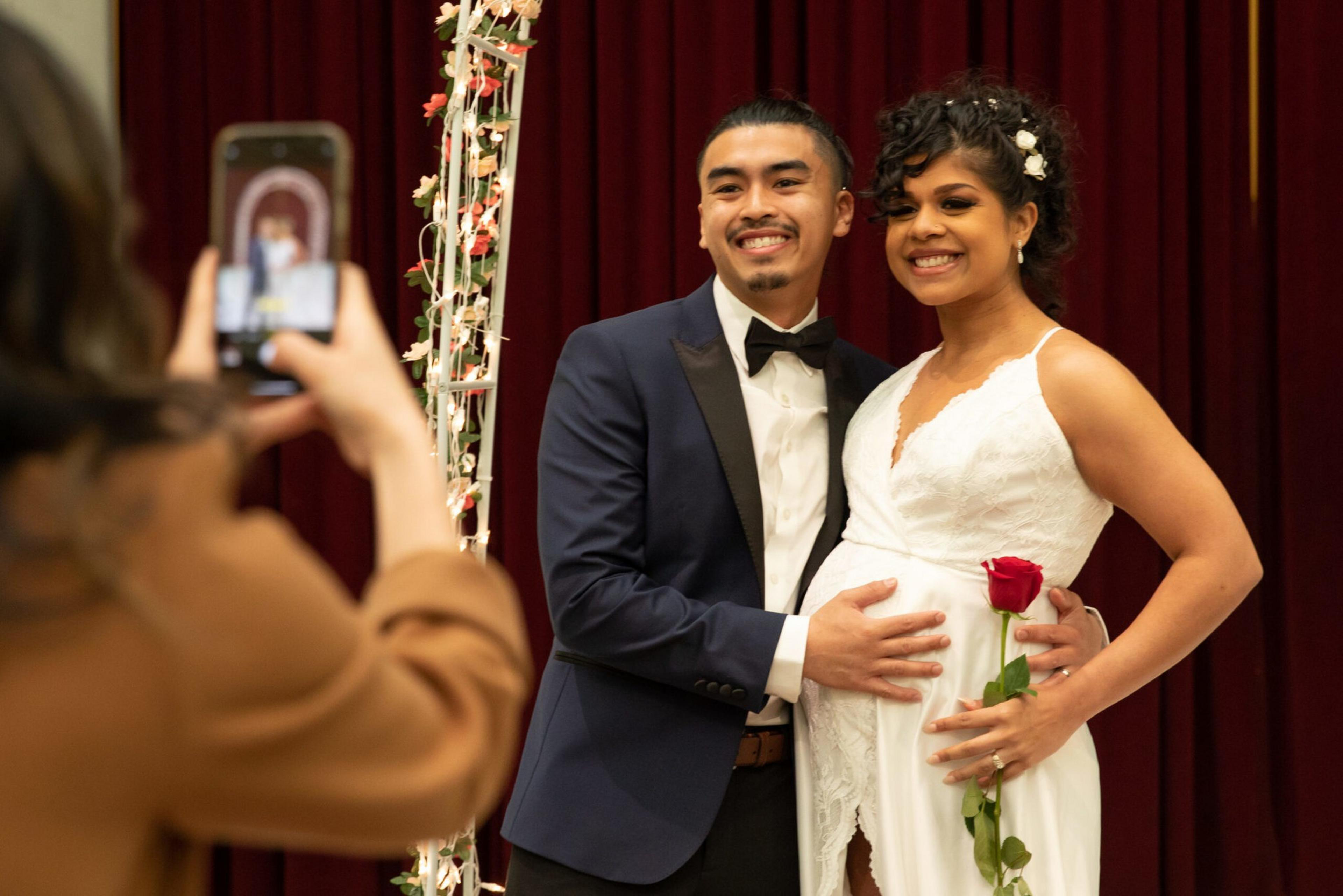A smiling couple poses for a photo; the woman in a white dress holds a red rose while the man in a tuxedo gently touches her belly, suggesting pregnancy.