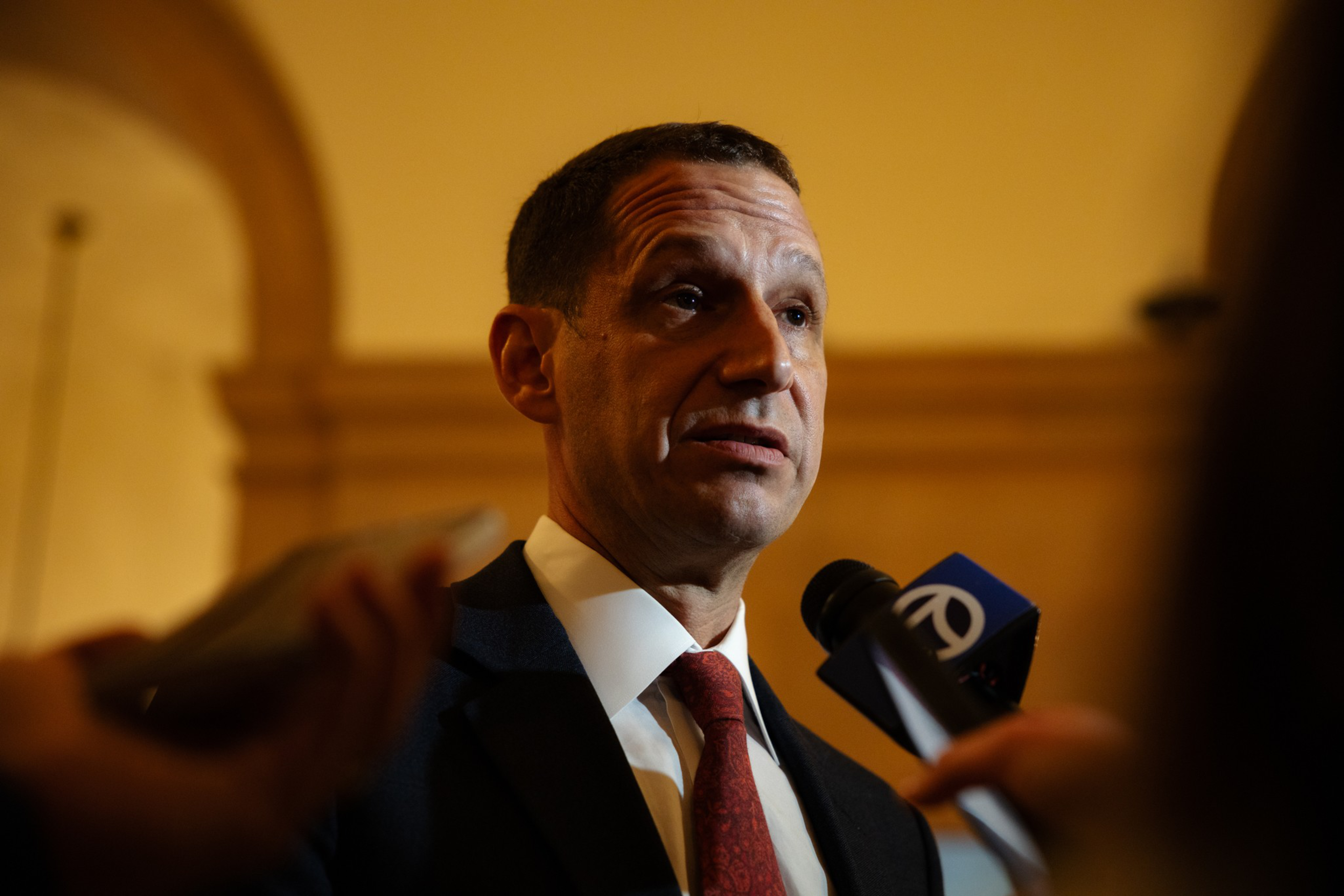 A man in a suit and red tie speaks into microphones, with a golden arch and warm lighting in the background.