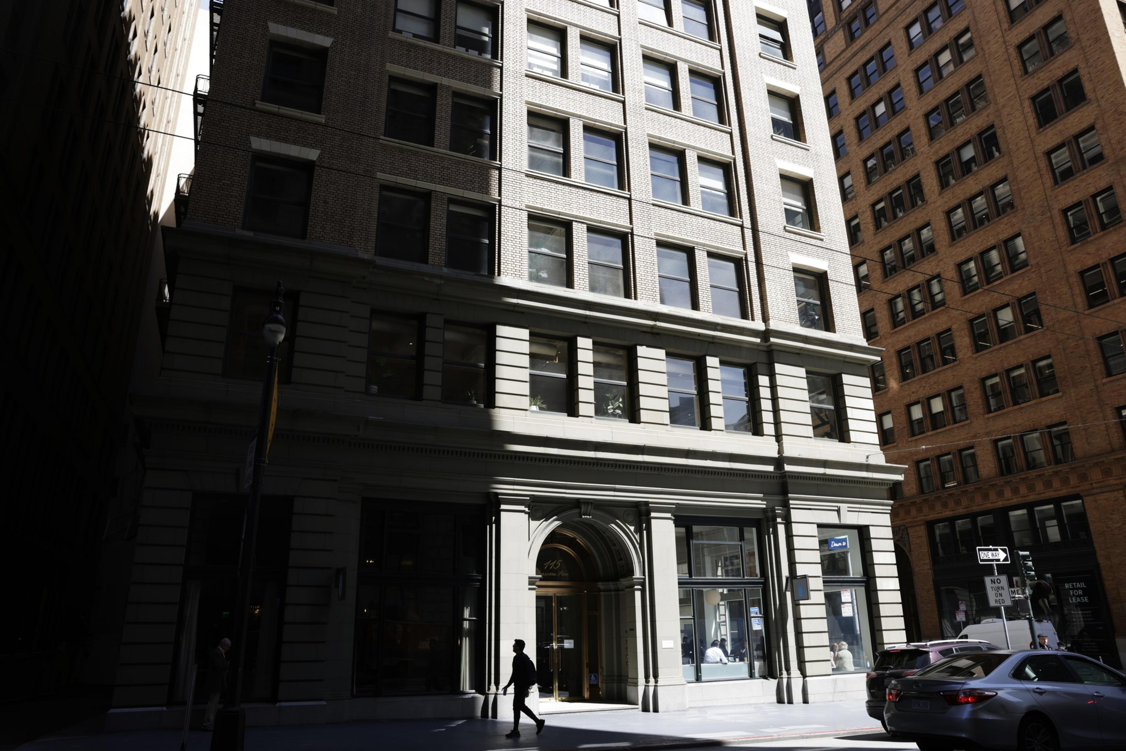A city street with tall buildings, shadowed pedestrians, and a car passing by.