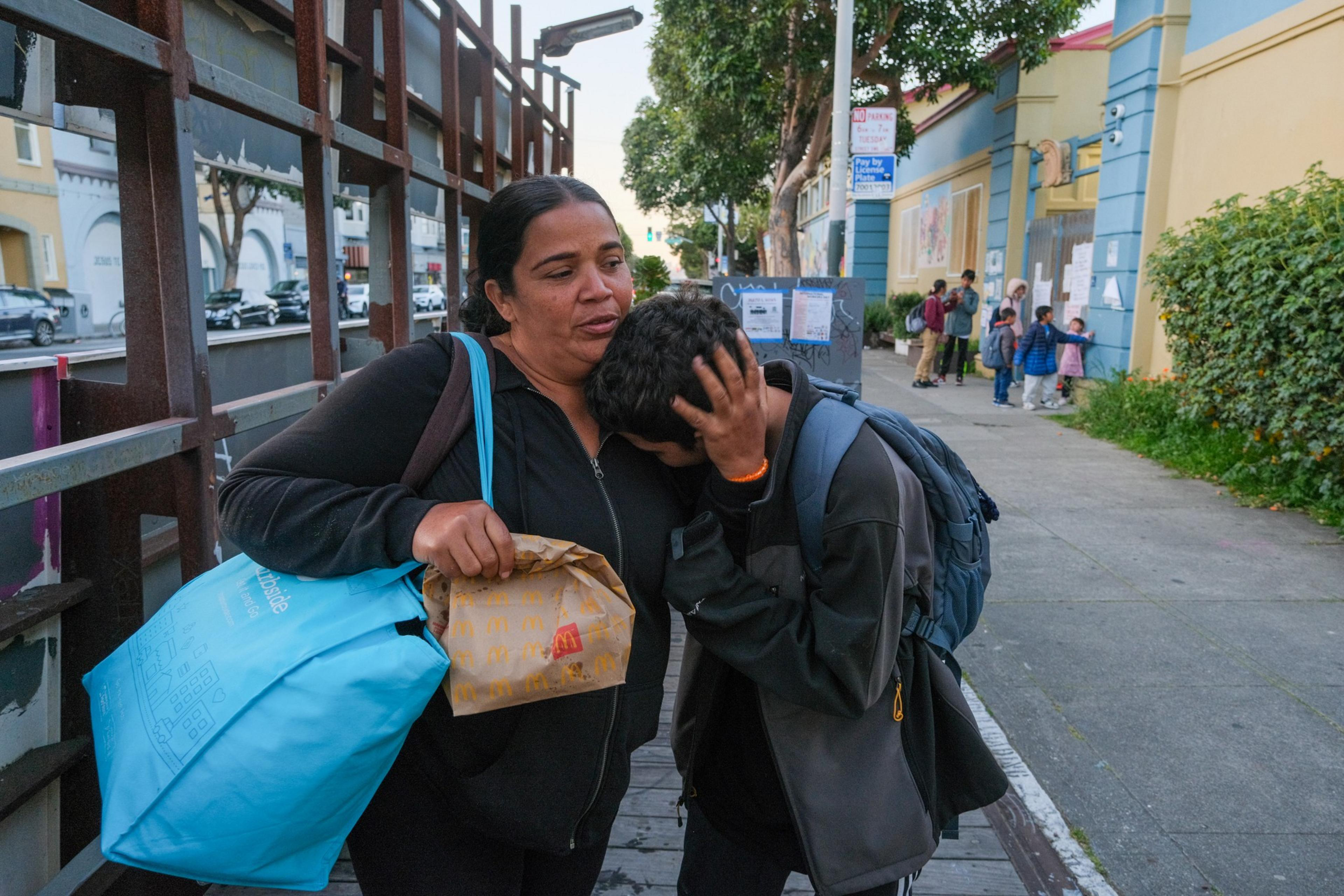 A woman comforts a young person on a sidewalk; both seem distressed, with a fast-food bag in view.