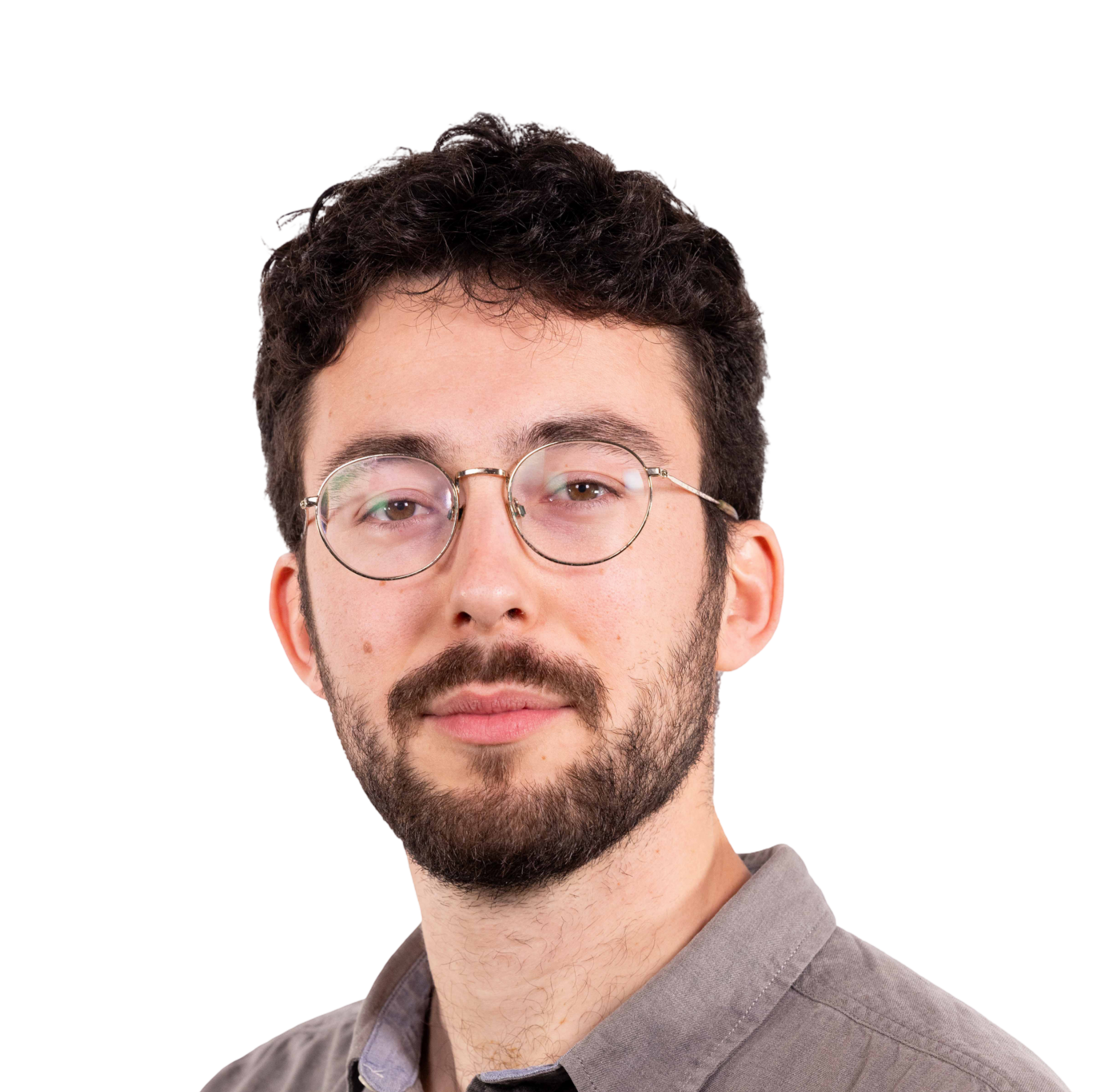 A young man with curly dark hair, beard, and round glasses wears a gray button-up shirt and looks confidently at the camera.