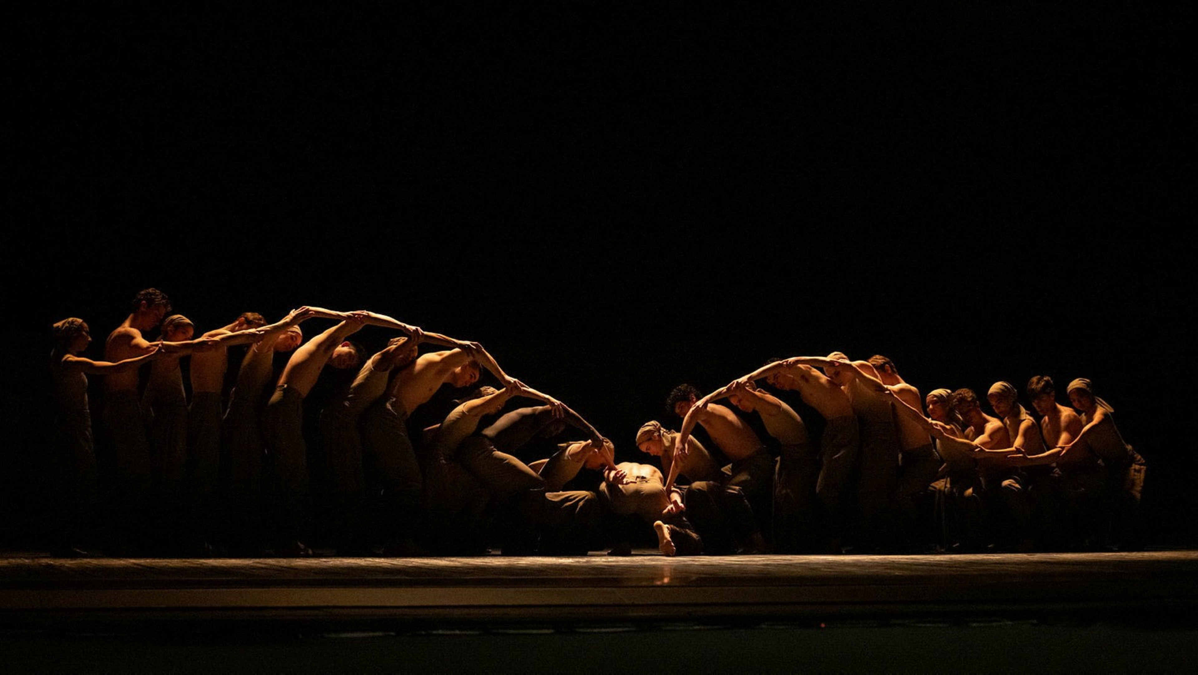 A group of dancers in brown costumes form an arch with their bodies on a dimly lit stage, with one person lying in the center underneath the arch.