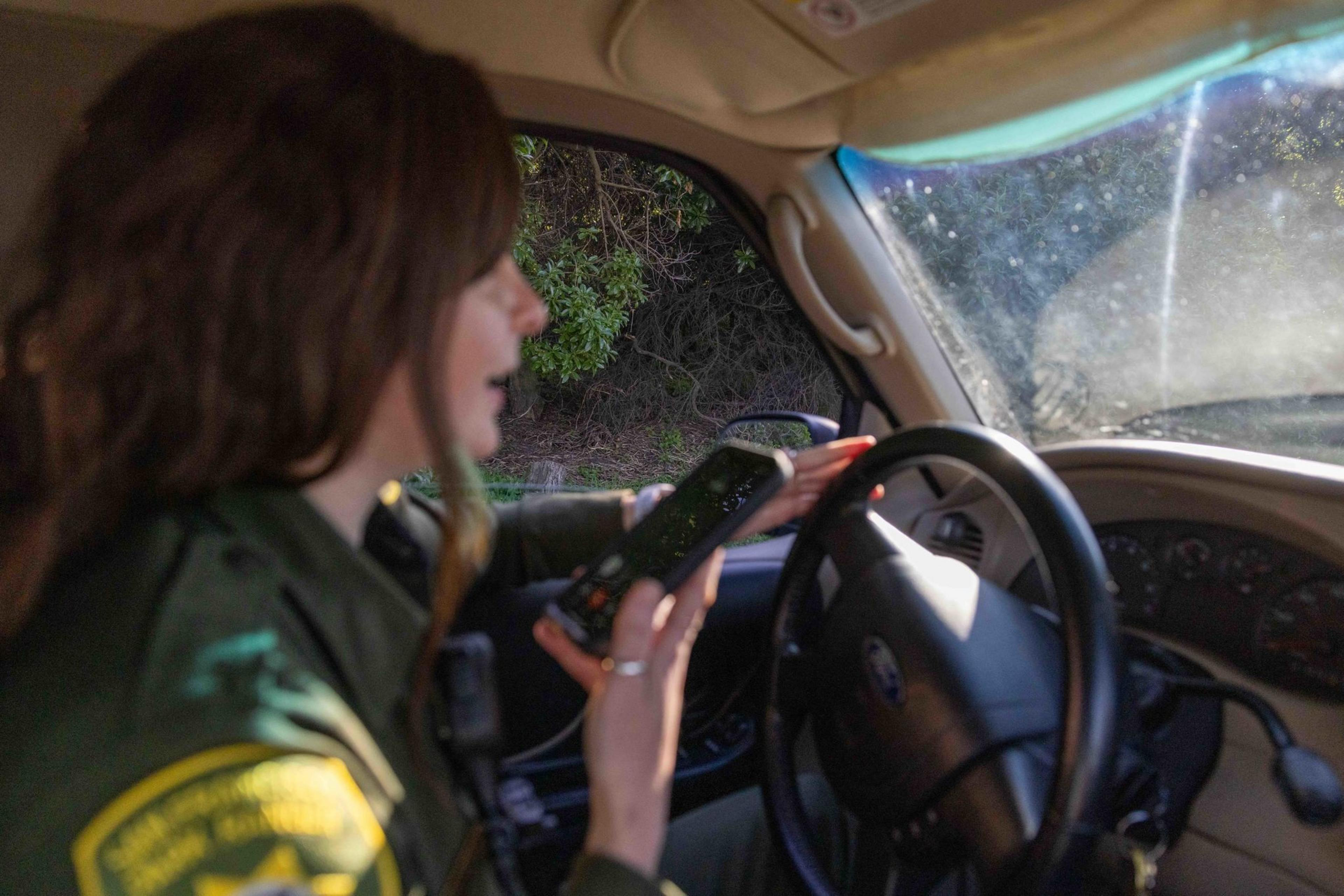 A person in a uniform is seated in a car, holding a smartphone and speaking. Sunlight illuminates the interior, and foliage is visible through the window.