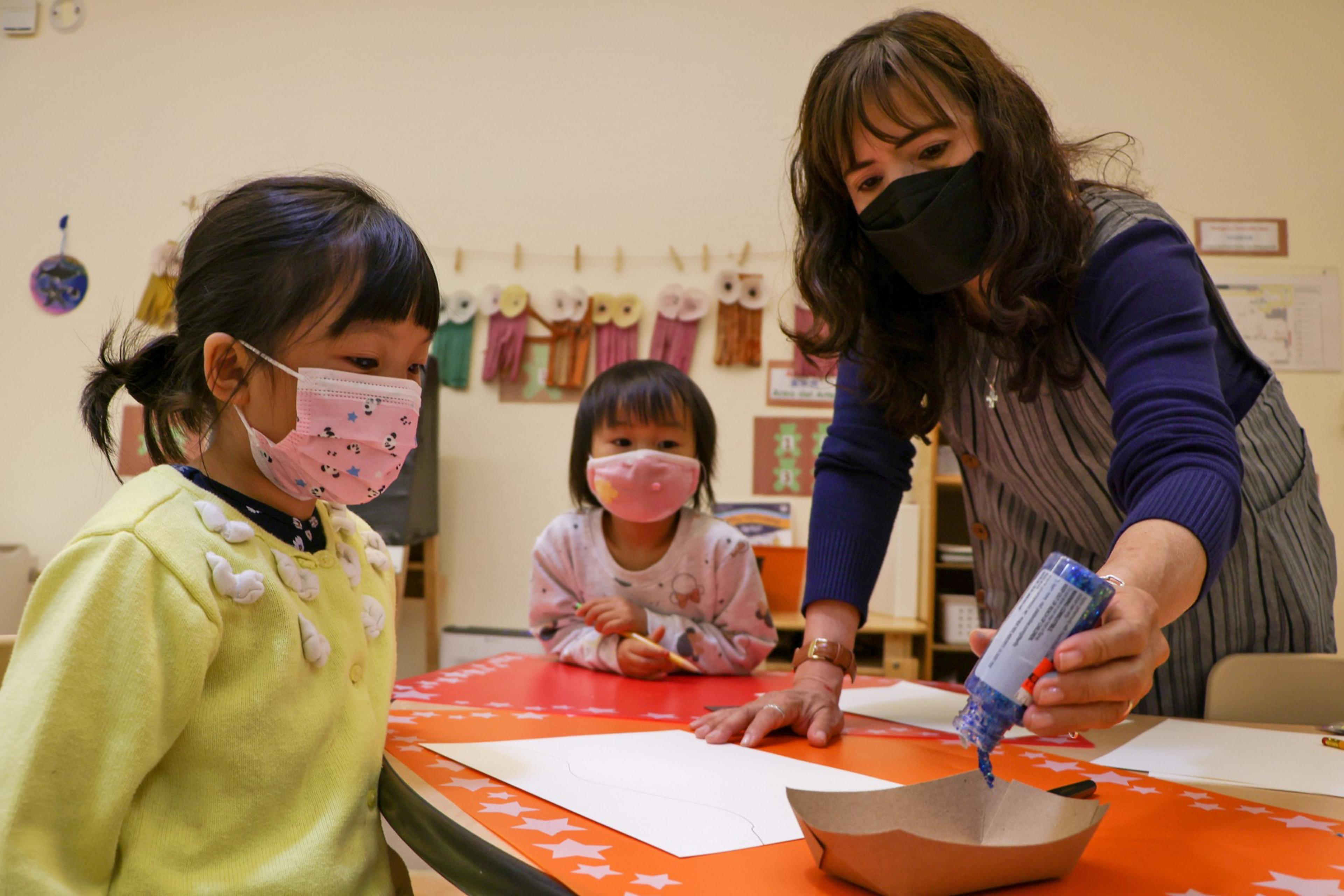 A woman wearing a black mask squeezes blue glue onto a tray while two young girls in pink masks watch attentively at a bright orange table.