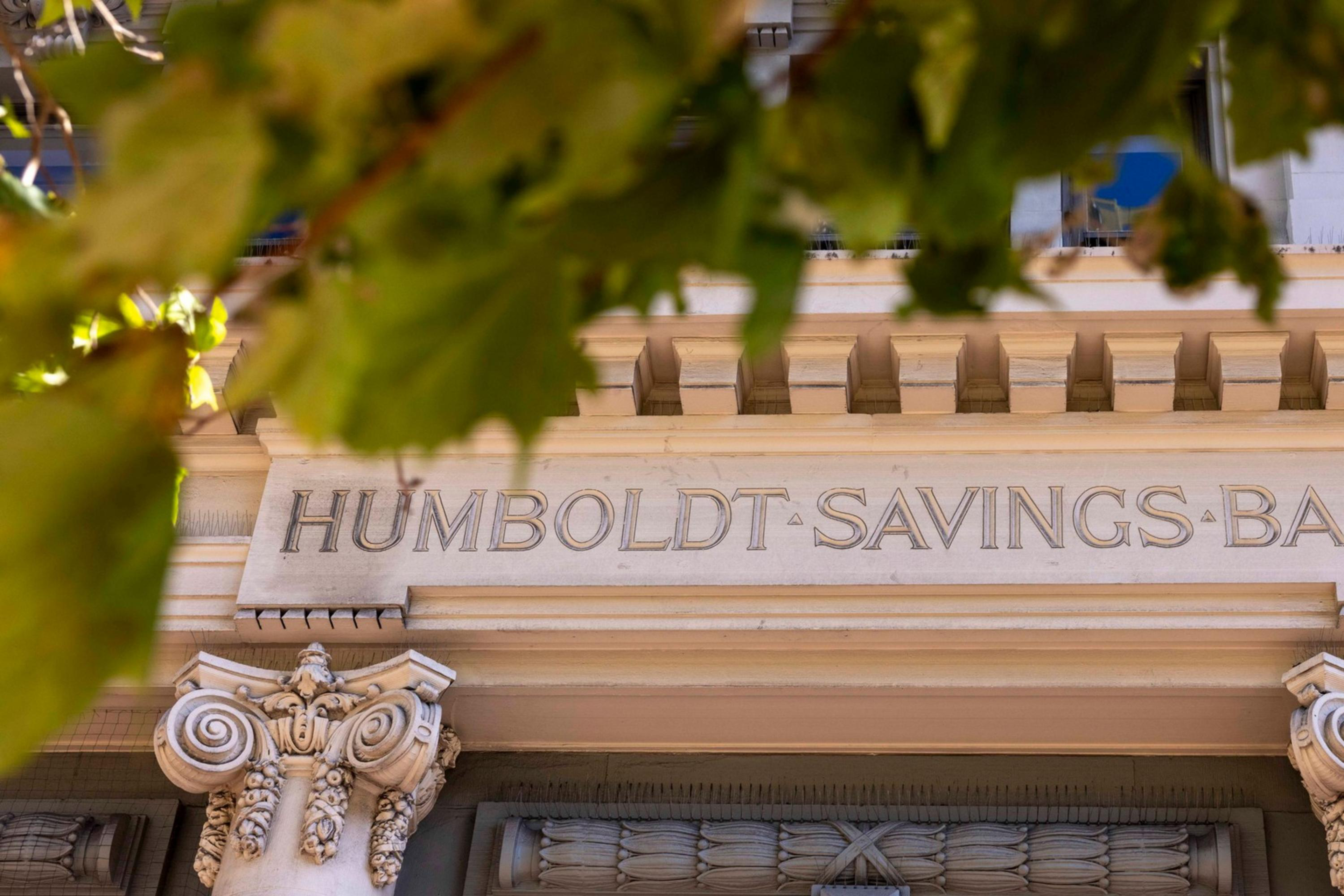 The image shows the facade of Humboldt Savings Bank, featuring classical architectural elements like ornate columns and intricate stone detailing, partially framed by tree leaves.