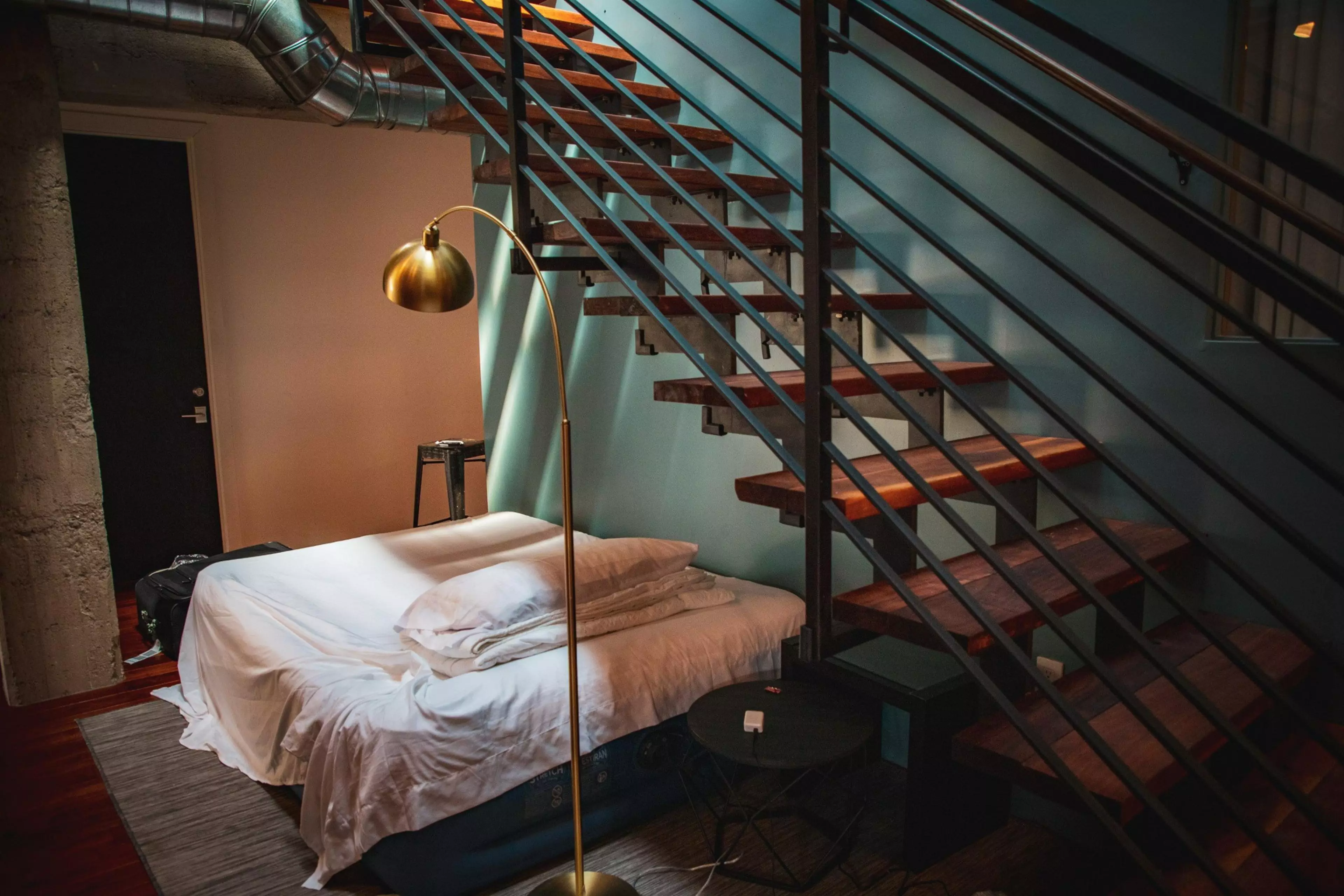 The image shows a loft-style room with a bed under wooden stairs, a gold floor lamp, and a small black table. The background includes exposed pipes and a door.