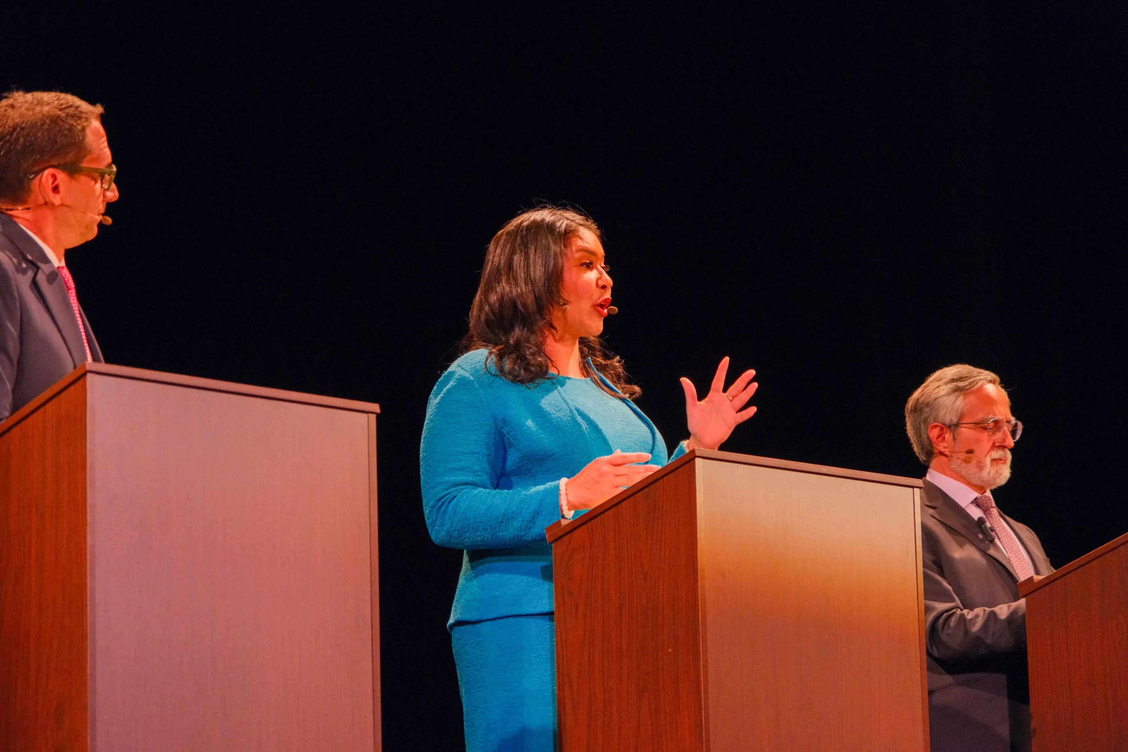 Three individuals stand at podiums on a dimly lit stage. The woman in the middle speaks with a hand raised, while the two men on either side listen intently.