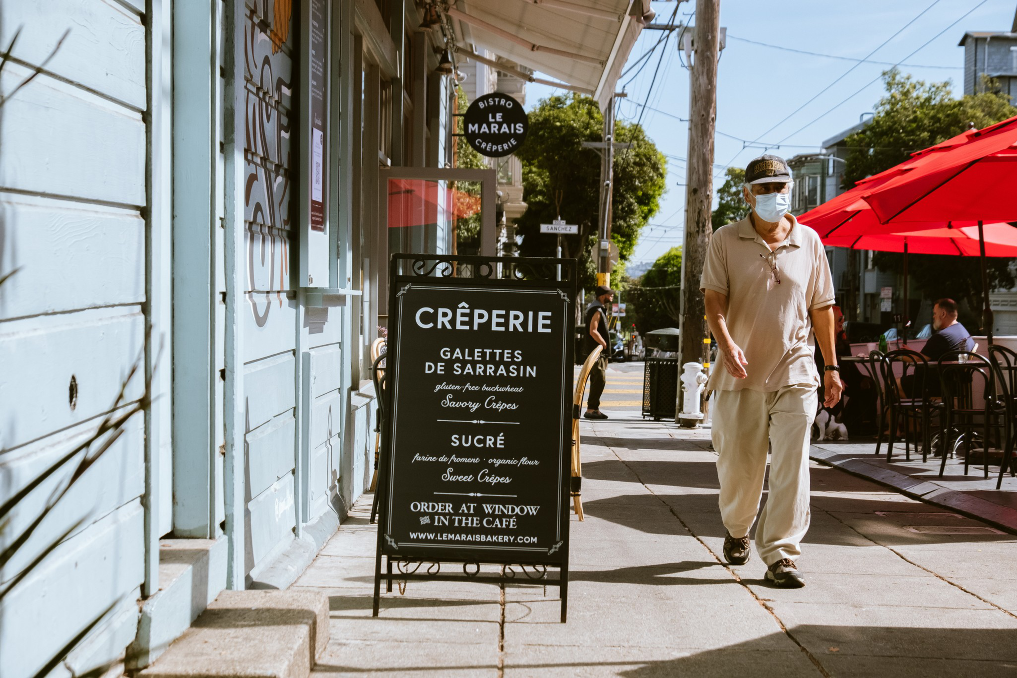 A sidewalk with a man walking past a crêperie sign, outdoor seating under red umbrellas in the background.