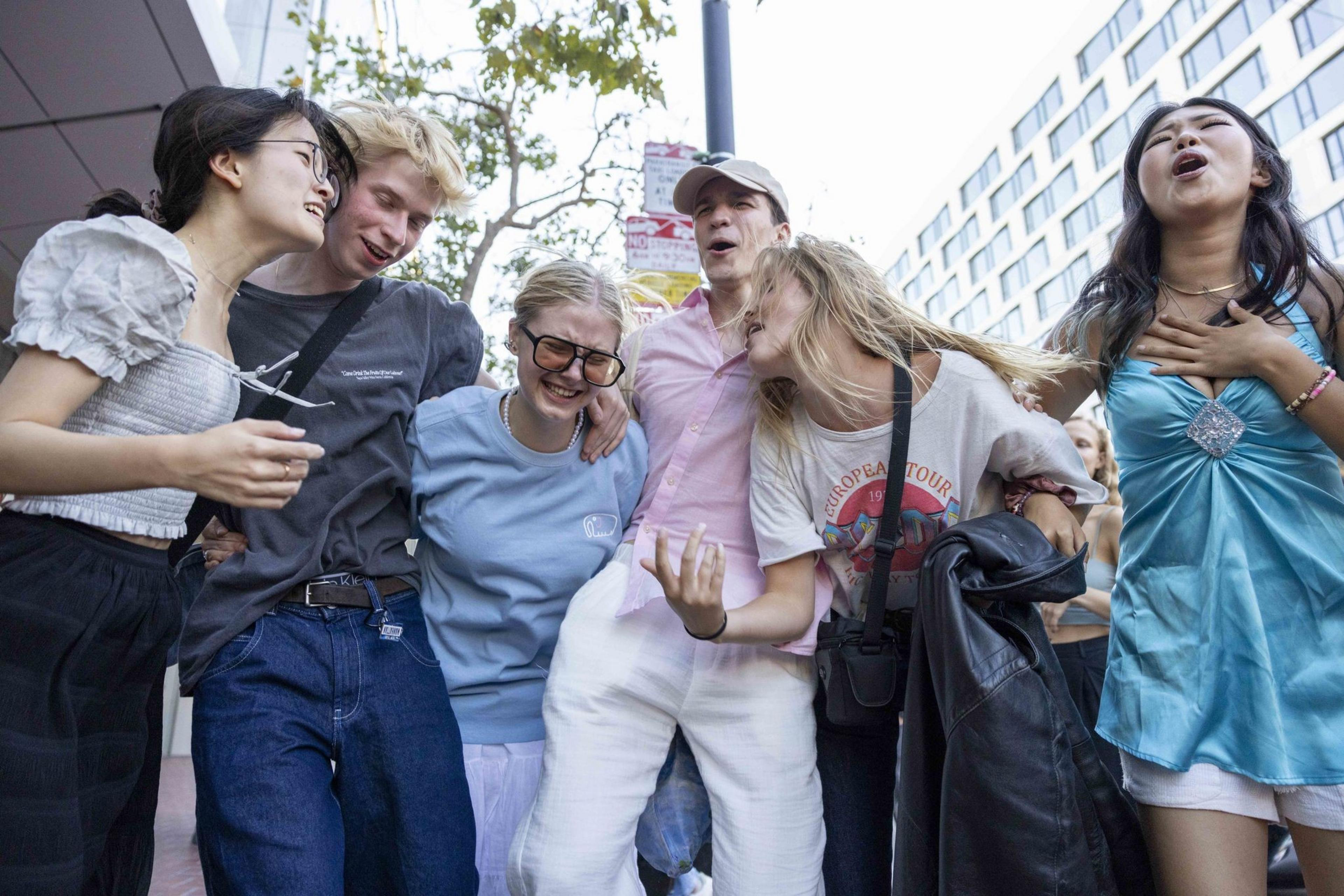 Six friends are laughing, singing, and embracing each other outdoors on a sunny day, creating a joyful and energetic atmosphere.