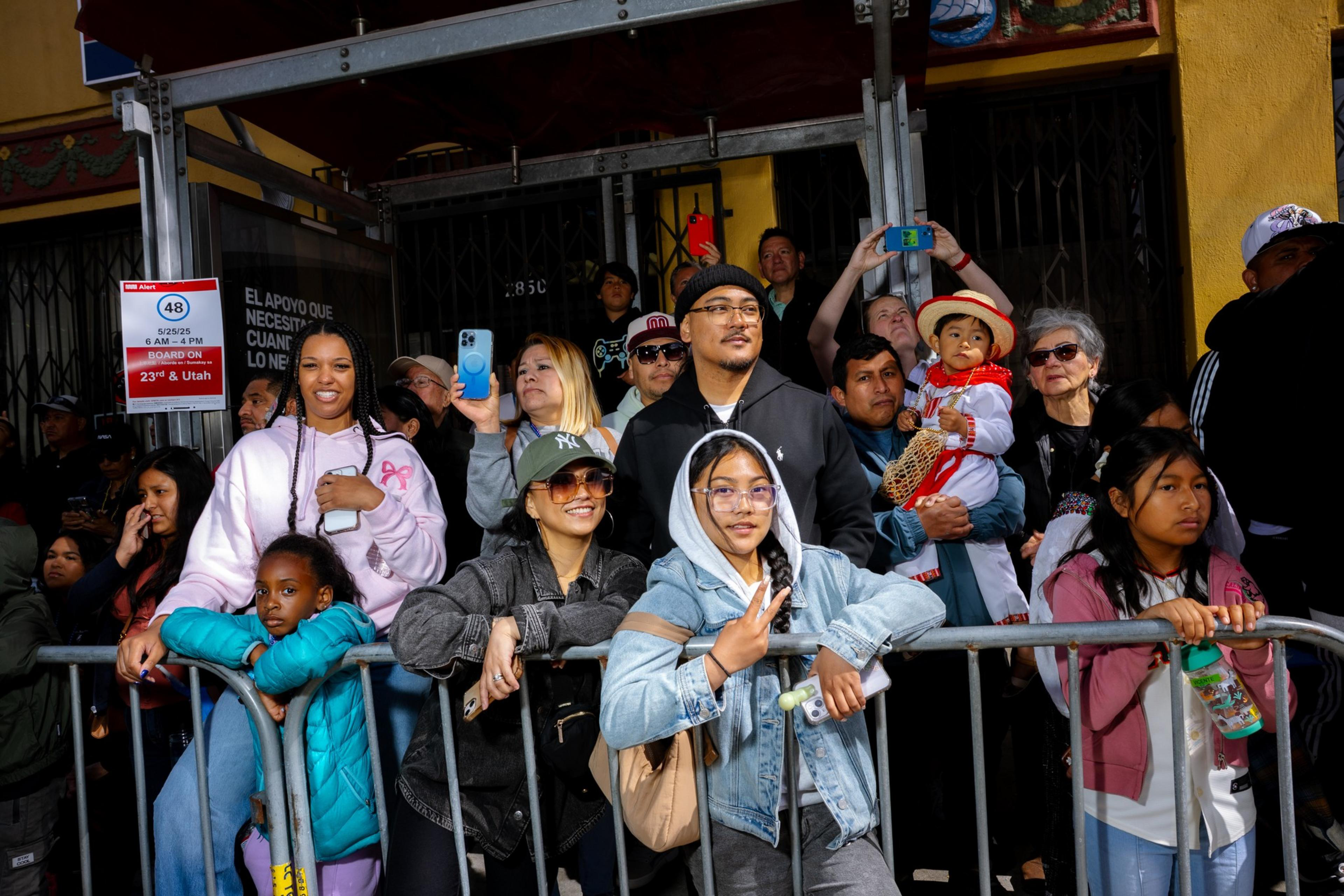 A diverse group of people stands behind a metal barrier, some smiling and holding phones. A child in a colorful outfit is held by a man, attracting attention.