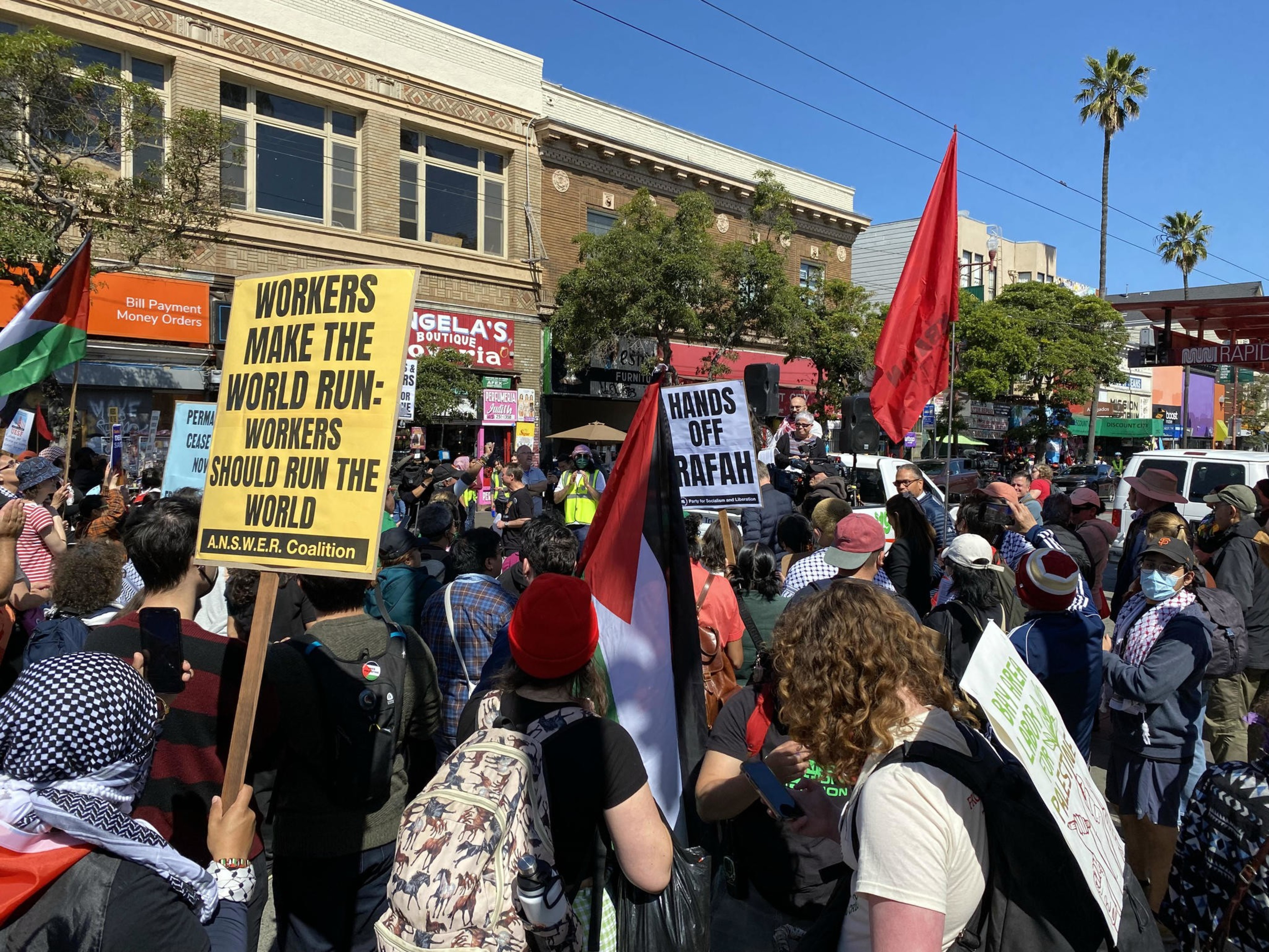 A crowd of protesters with signs, one reading &quot;WORKERS MAKE THE WORLD RUN,&quot; in an urban setting under a clear sky.