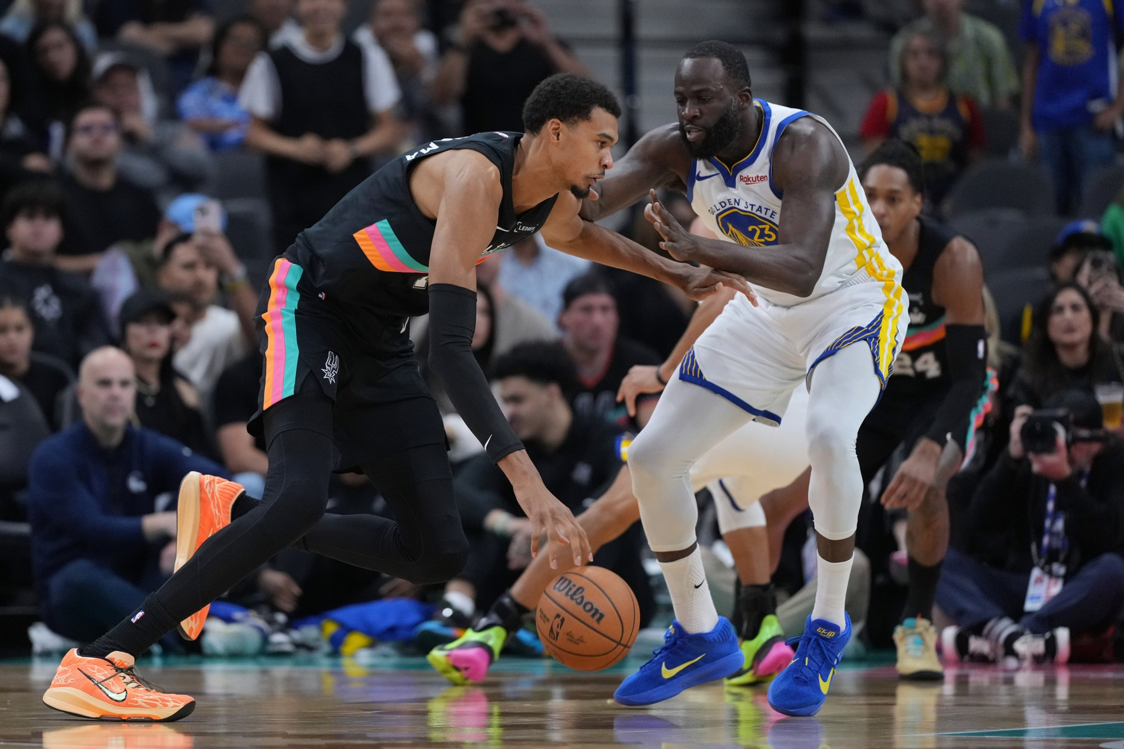 A basketball player in a black uniform dribbles the ball while being closely defended by a player in a white Golden State Warriors uniform.