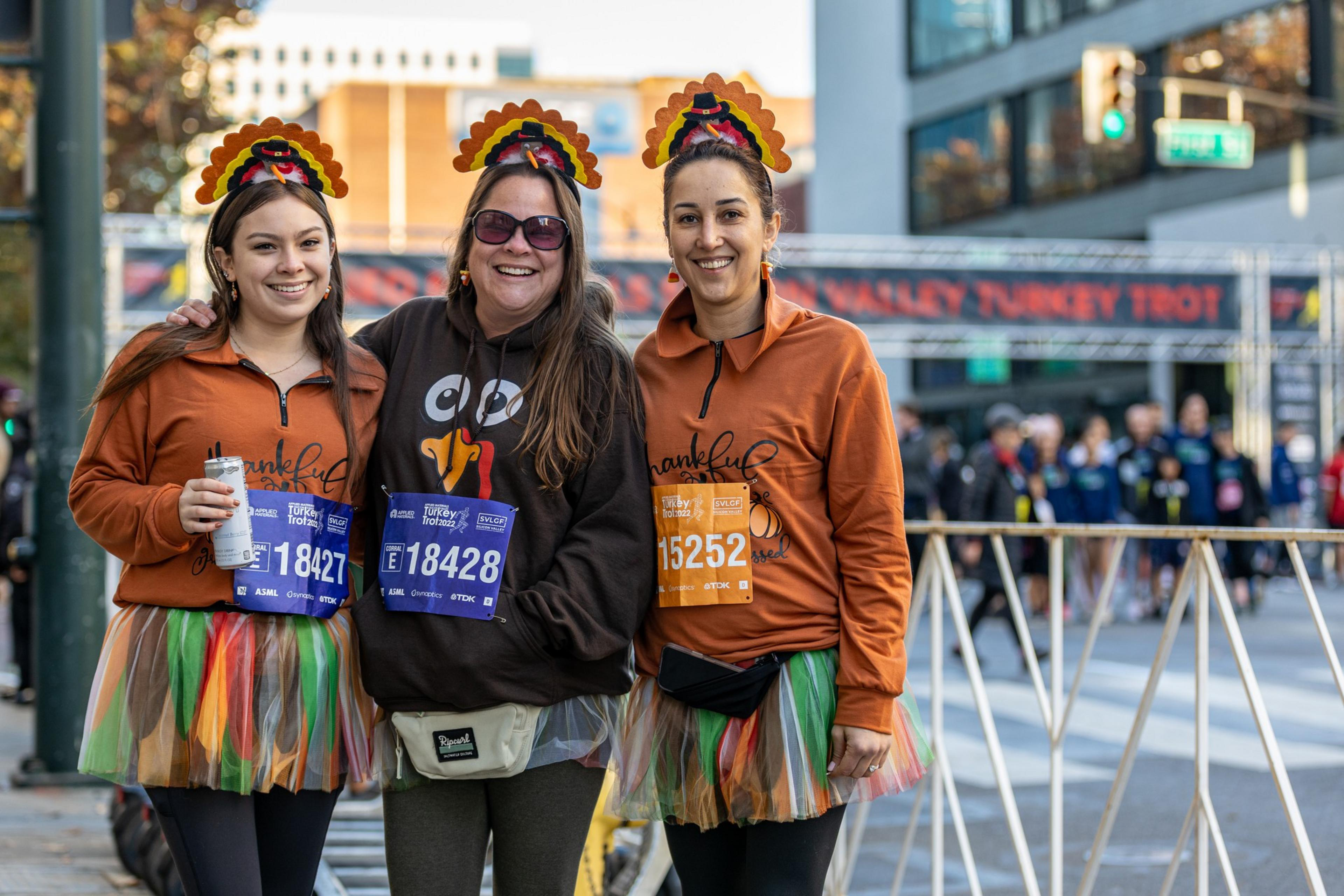 Three women stand at a race course wearing turkey crowns on their heads.