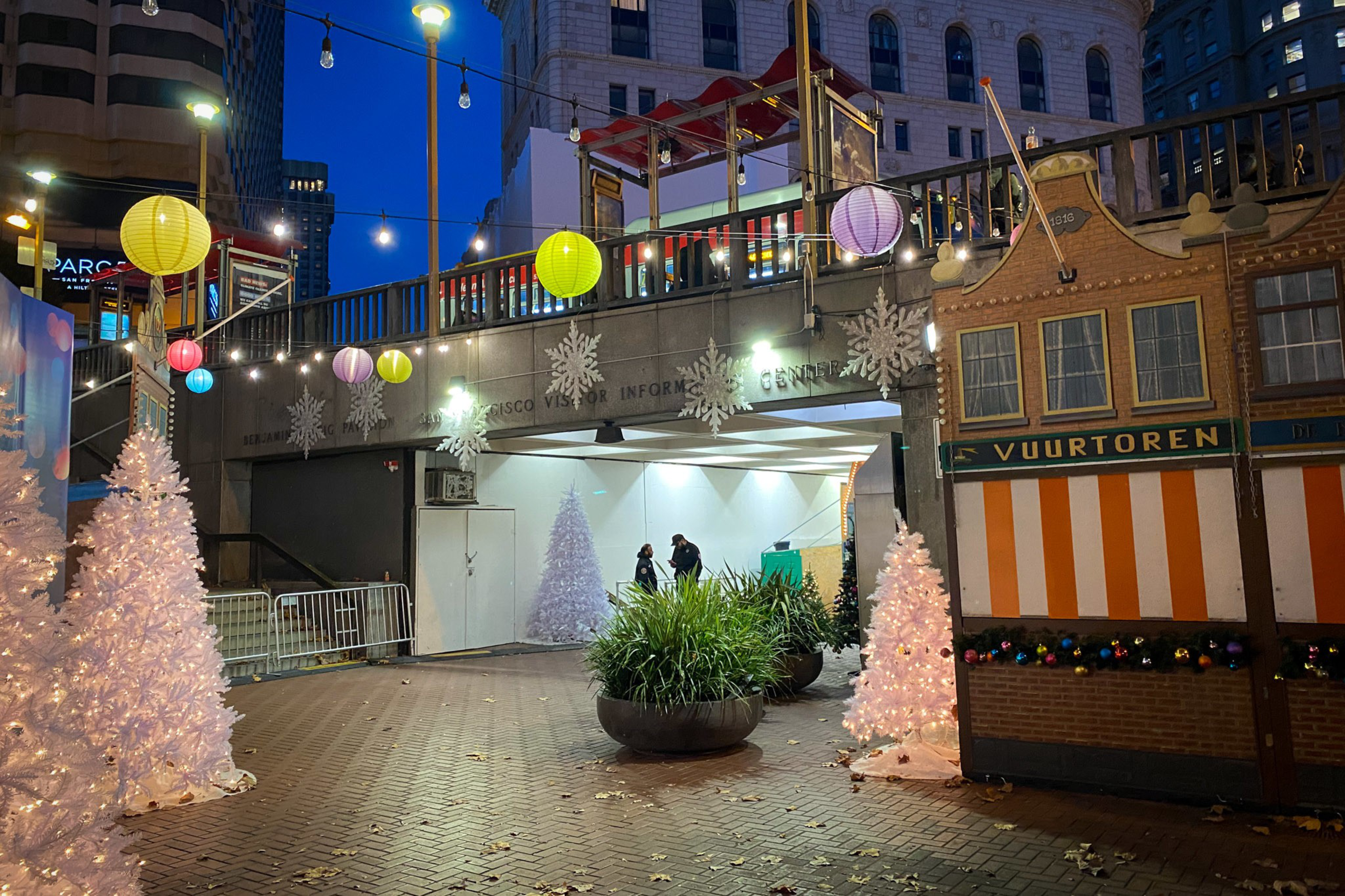White Christmas trees, colorful lanterns, and string lights decorate a city walkway near an information center at dusk, with people and plants nearby.
