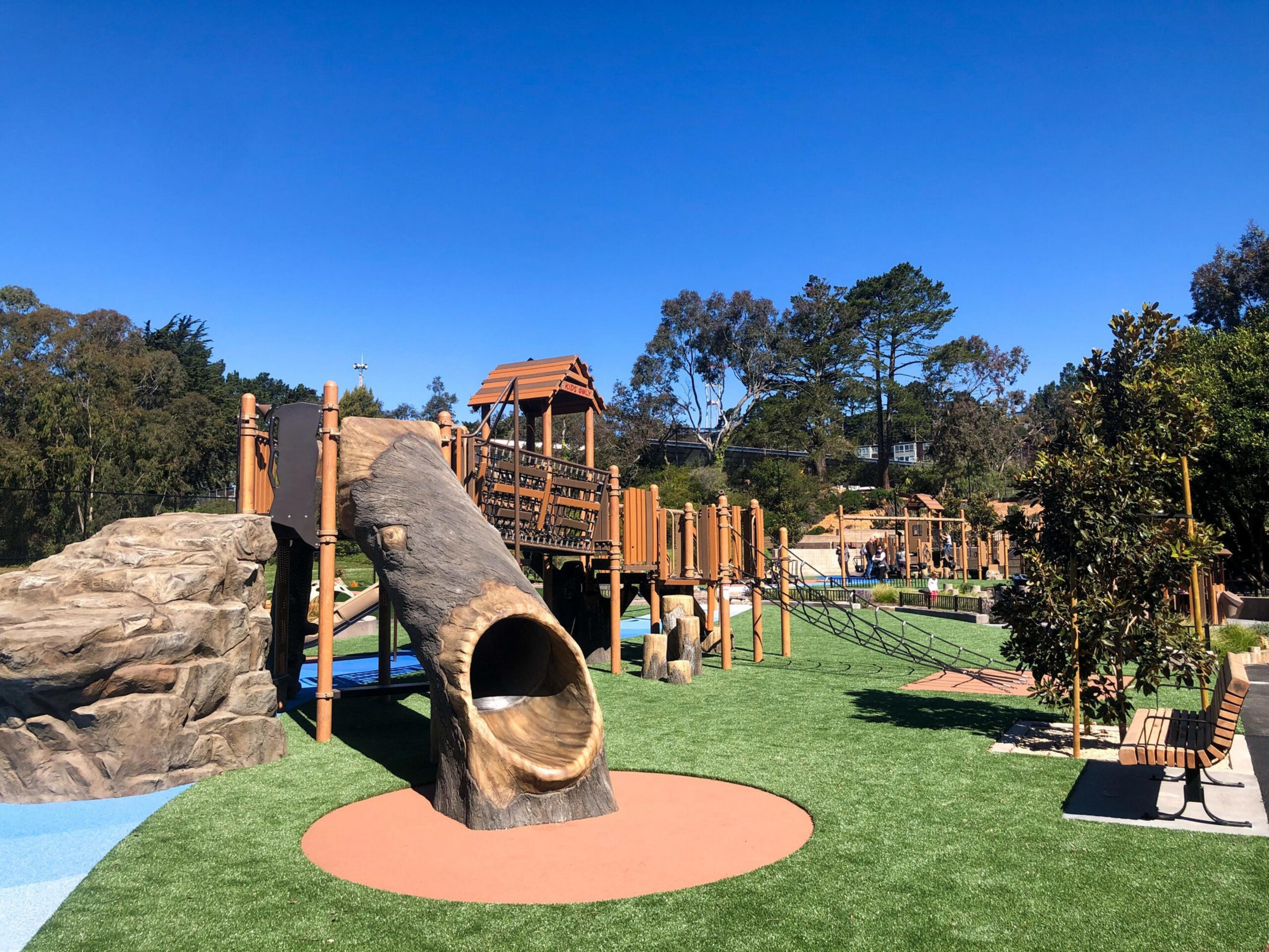 A playground with slide shaped like a hollow log, climbing structures, green artificial grass, benches, and trees under a clear blue sky.