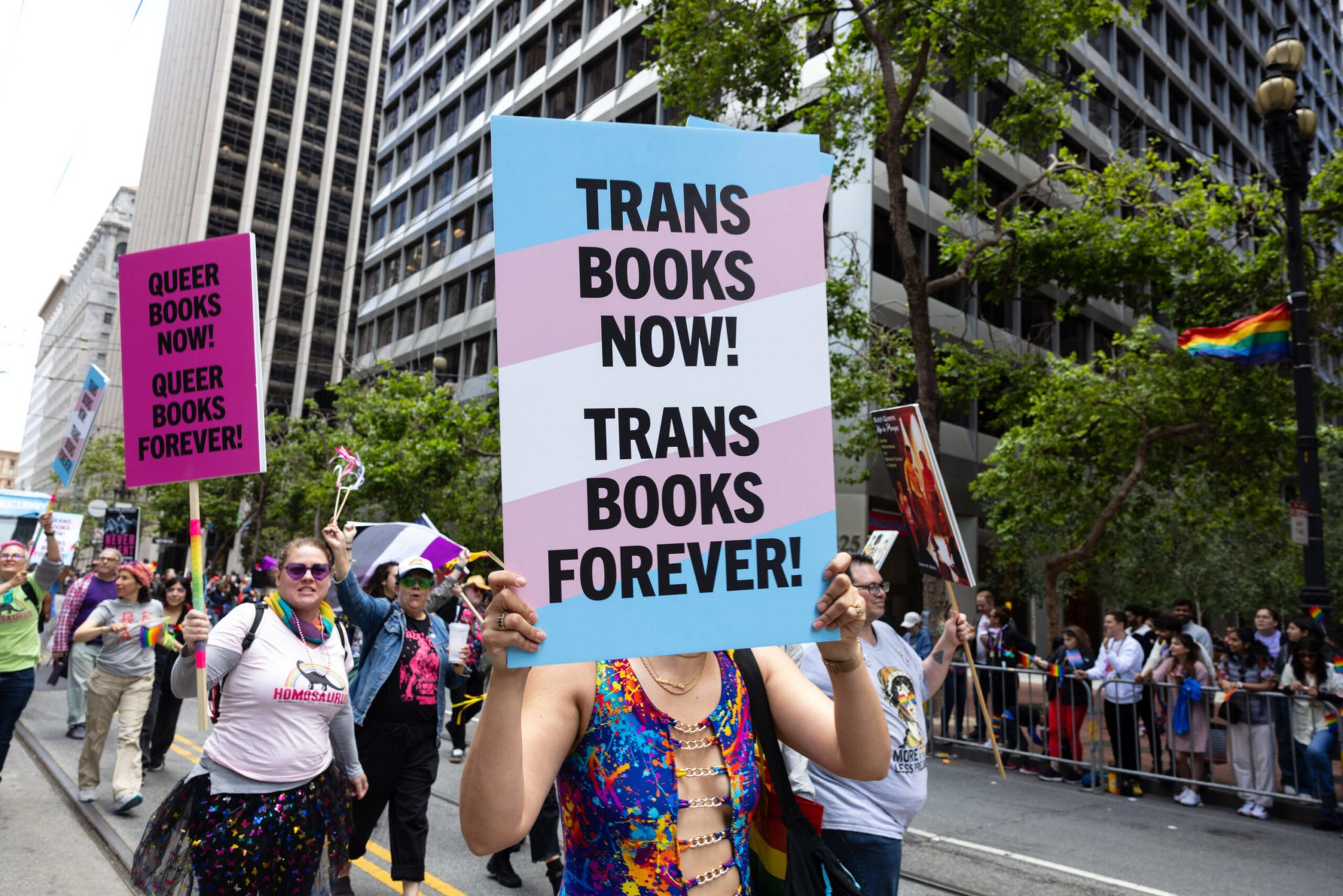 People march in a city street holding signs that say "TRANS BOOKS NOW! TRANS BOOKS FOREVER!" and "QUEER BOOKS NOW! QUEER BOOKS FOREVER!"
