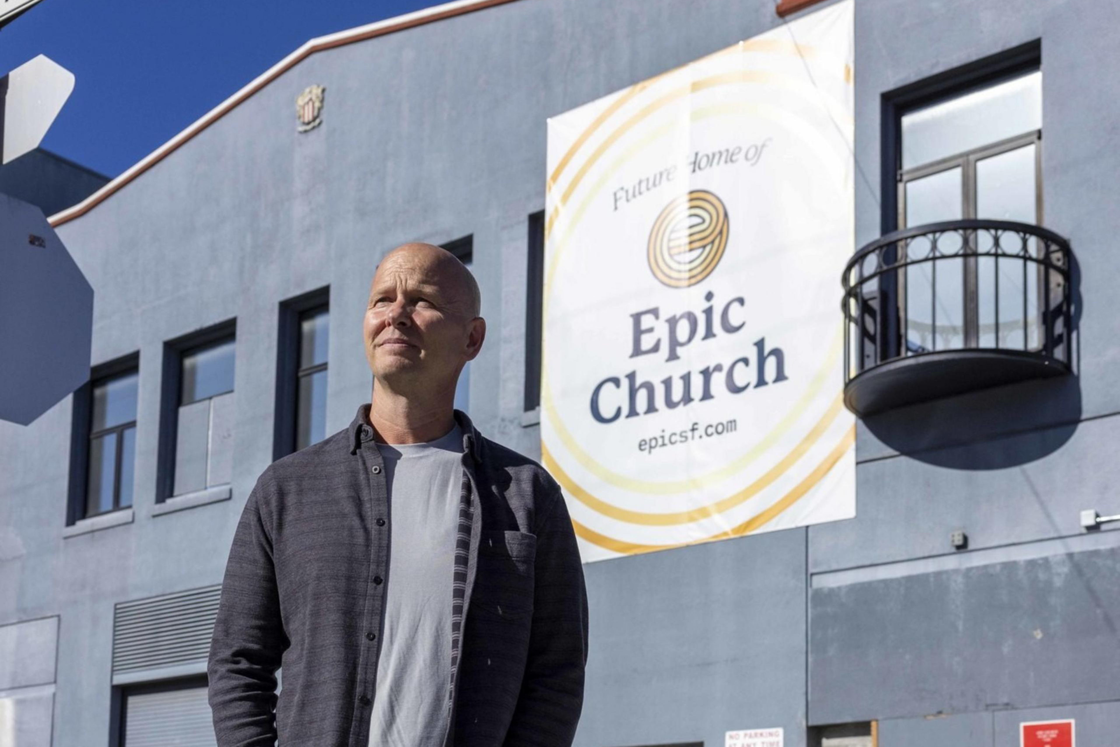 A person stands on a street corner in front of a blue building with a sign reading "Future Home of Epic Church" and looks thoughtfully into the distance.