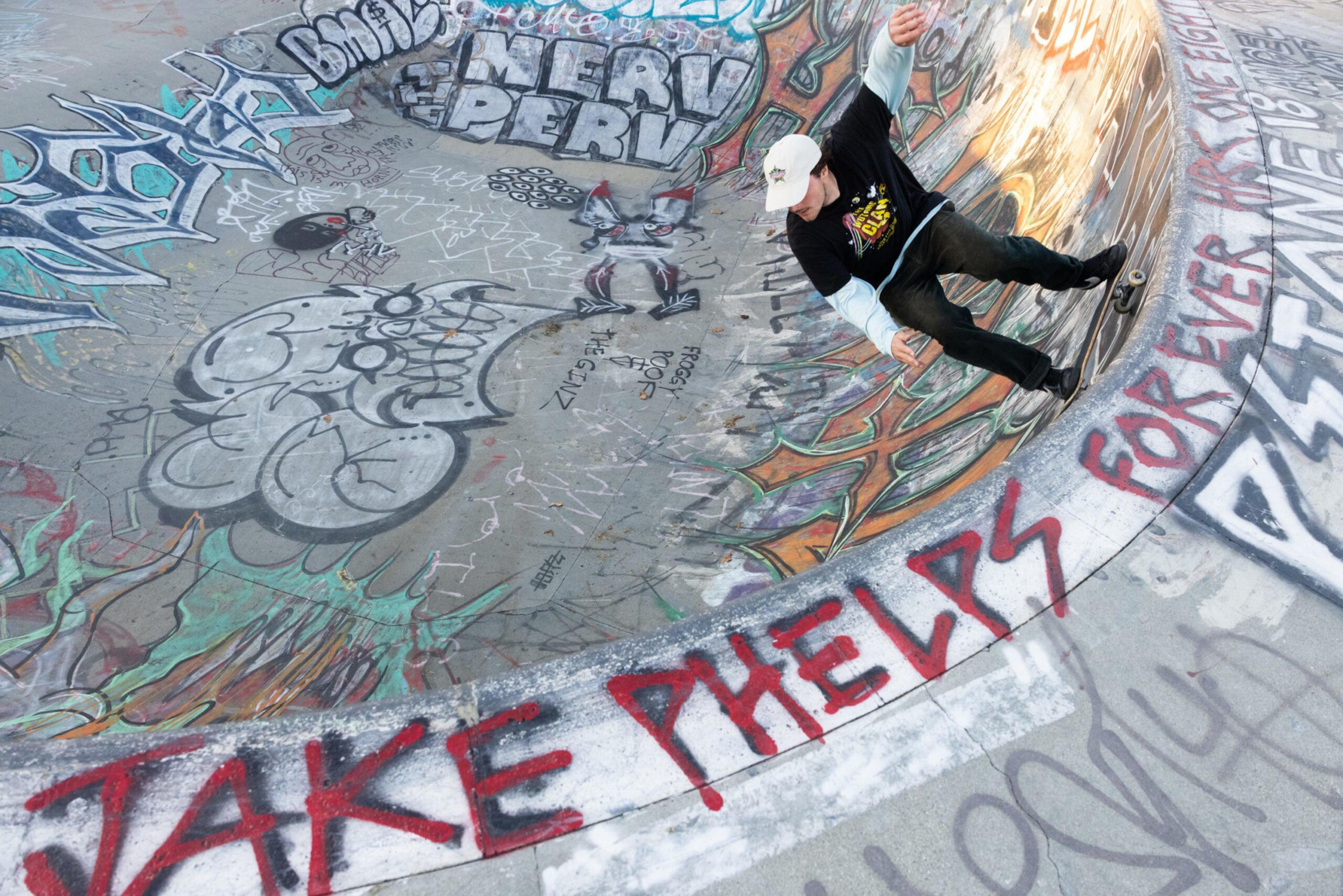 A skateboarder rides inside a graffiti-covered concrete bowl with vibrant drawings and the phrase "JAKE PHELPS FOREVER" spray-painted in red along the edge.