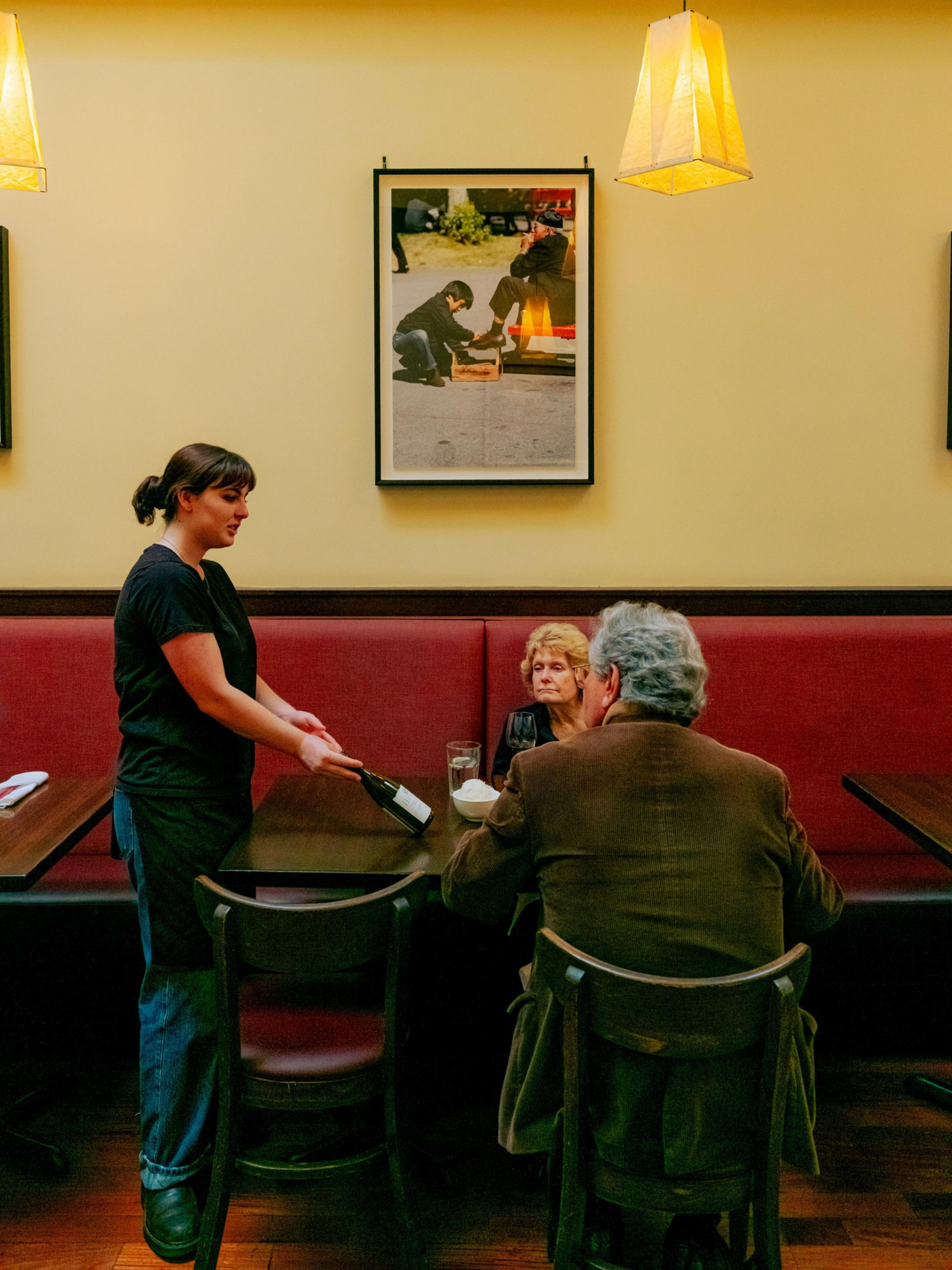 A waitress presents a wine bottle to a seated couple at a restaurant. A framed photo and warm light fixtures hang on a wall behind them.