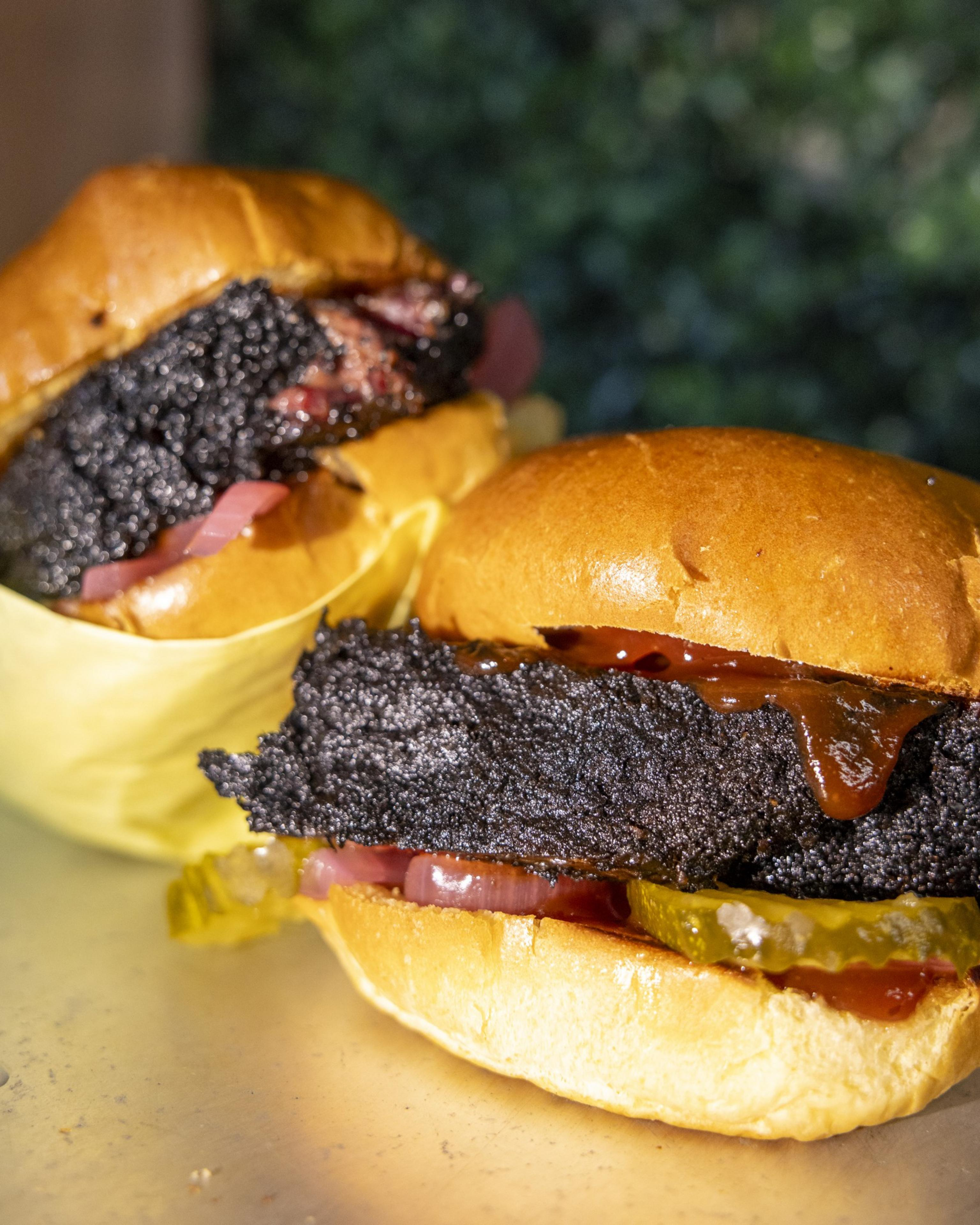 The image features two burgers with crispy, charred meat, pickles, onions, and sauce on shiny, golden buns on a light surface with a greenery background.