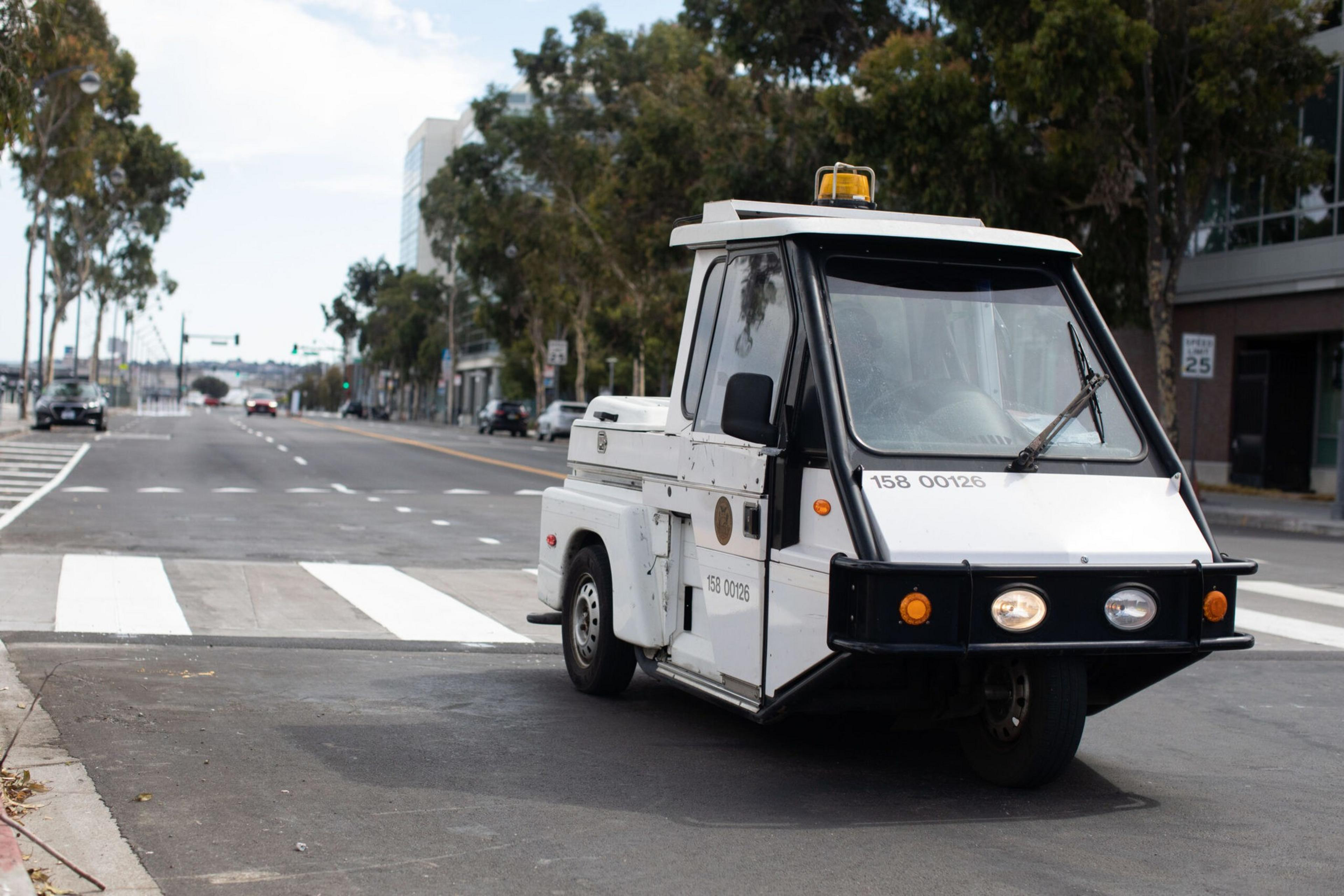 A small white three-wheeled utility vehicle with a single front headlight and a yellow beacon light on top drives on a city street near a crosswalk.