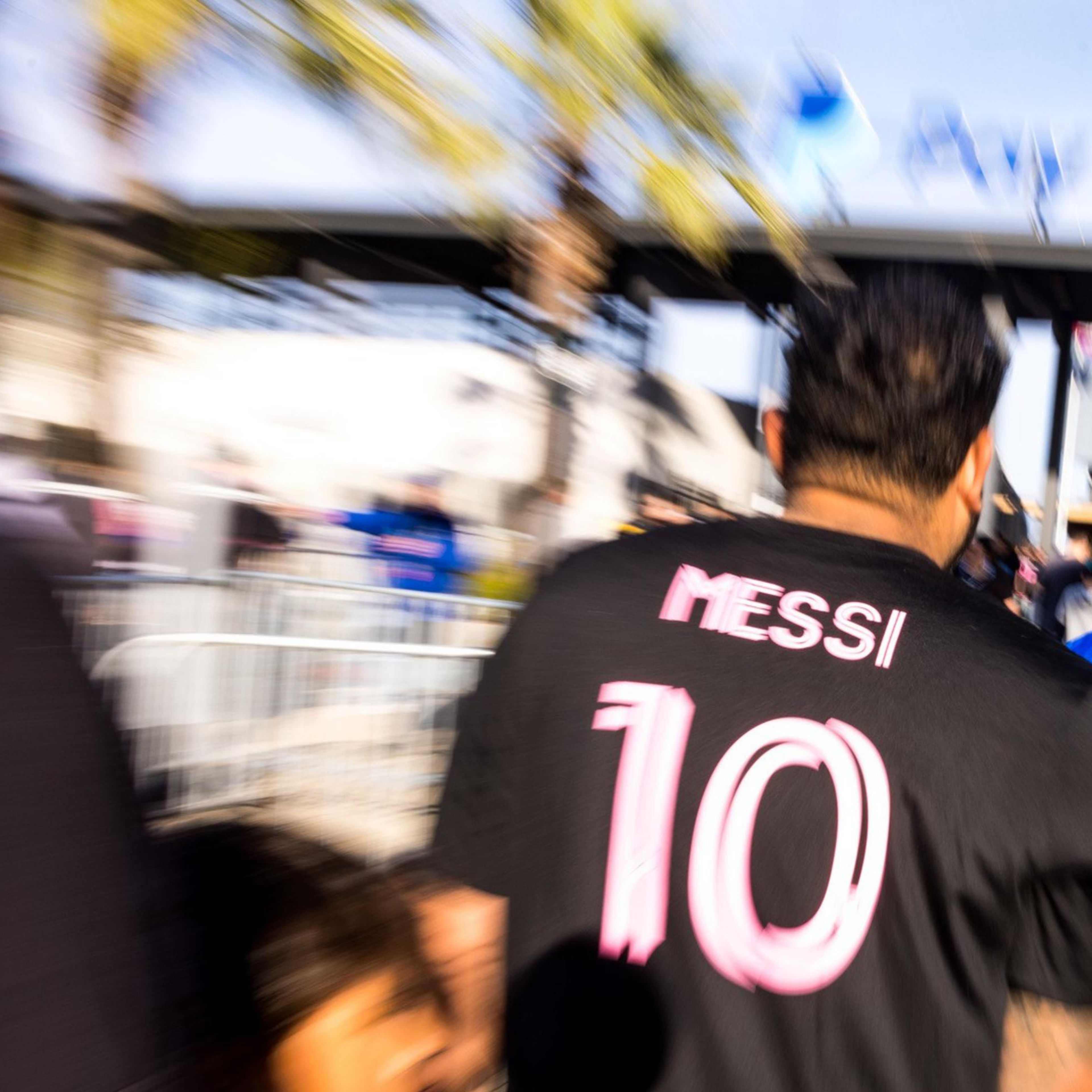 A crowd walks towards an entrance. One person in the foreground wears a black shirt with "MESSI 10" in pink letters. The image has a dynamic, blurred effect.