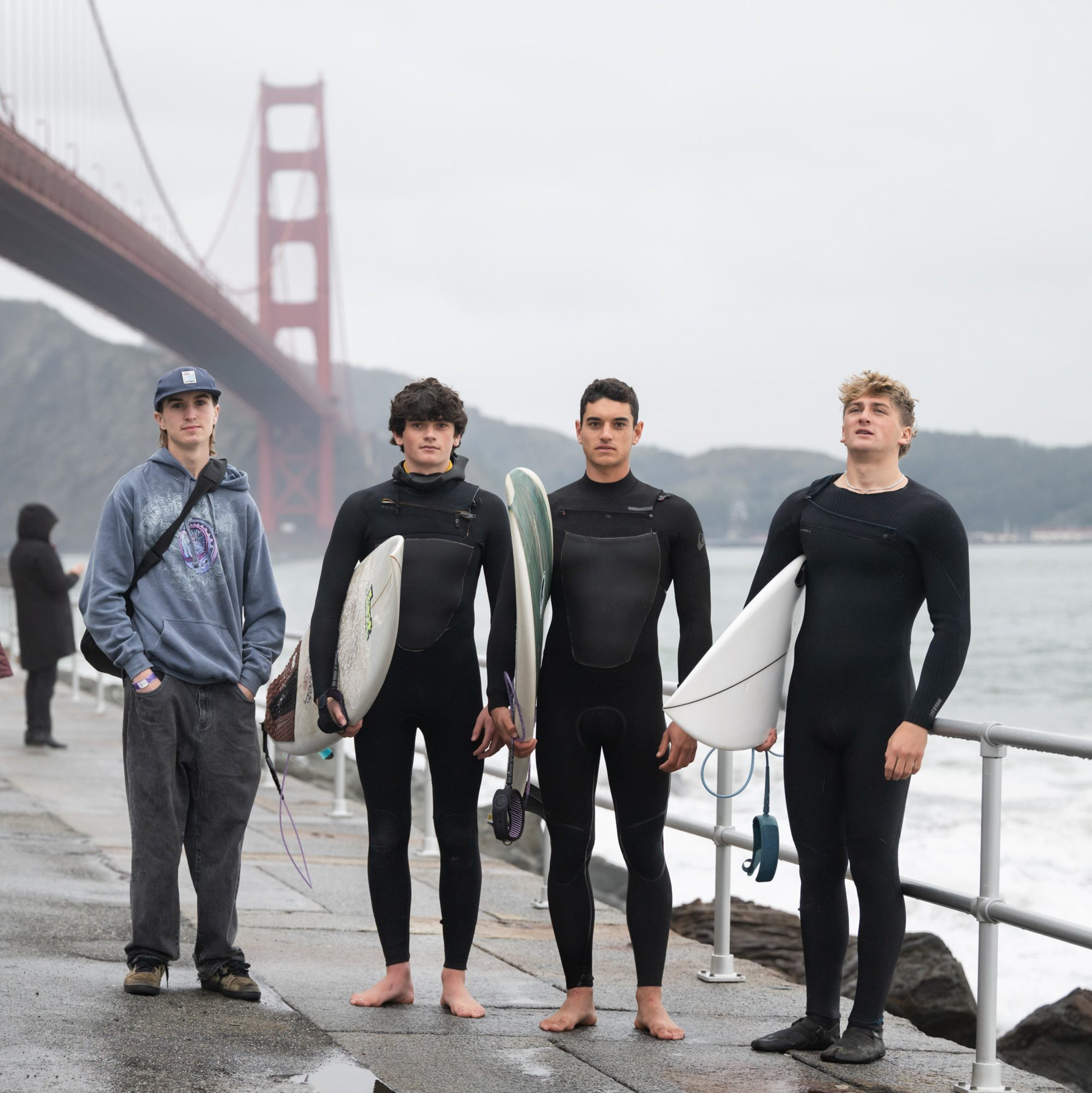 Four young men stand on a wet walkway near the ocean, three in wetsuits holding surfboards, and one in casual clothes. The Golden Gate Bridge is in the background.