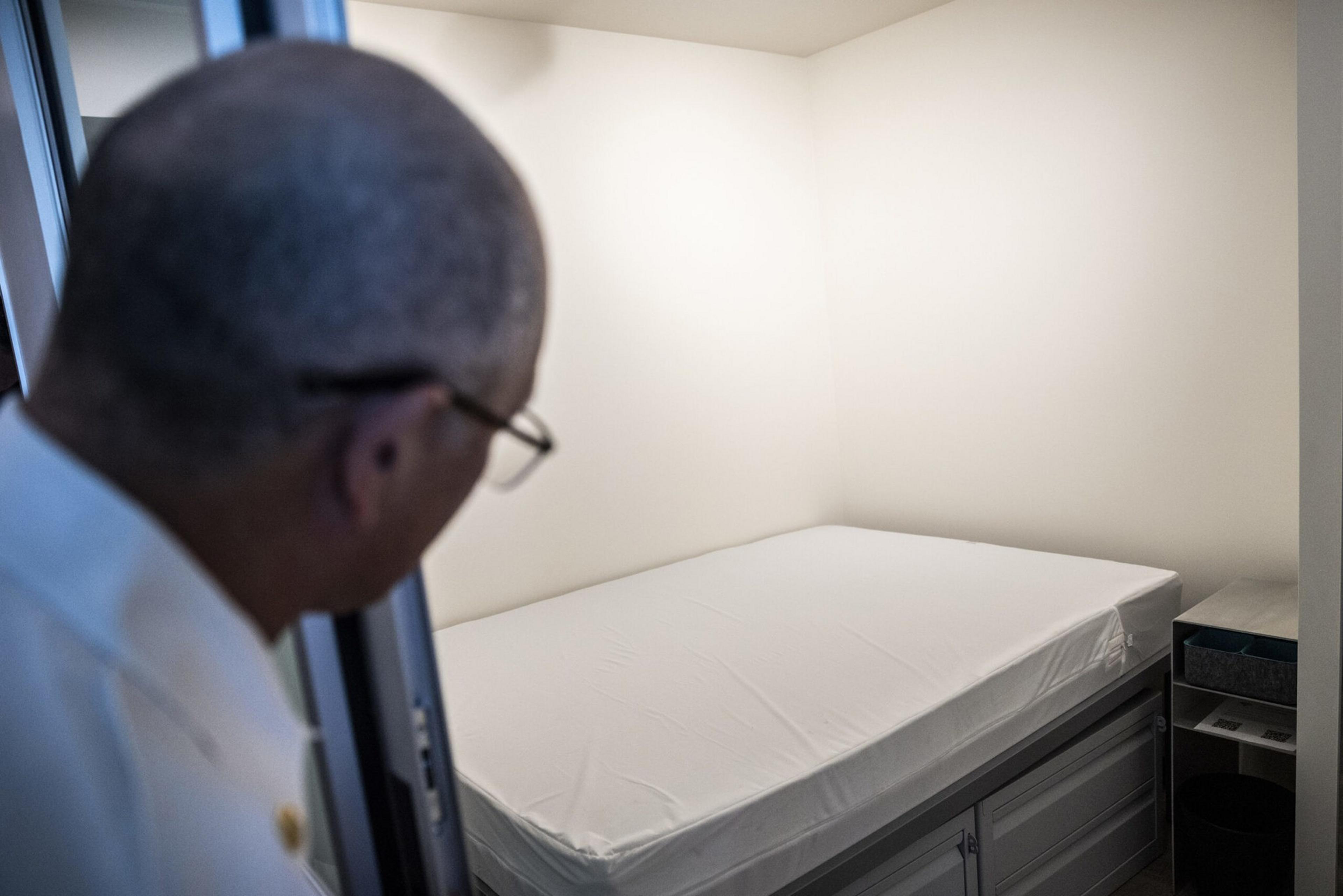 A man peers into a small, plain room with a single bed covered by a white sheet and a small bedside table with an electronic device.