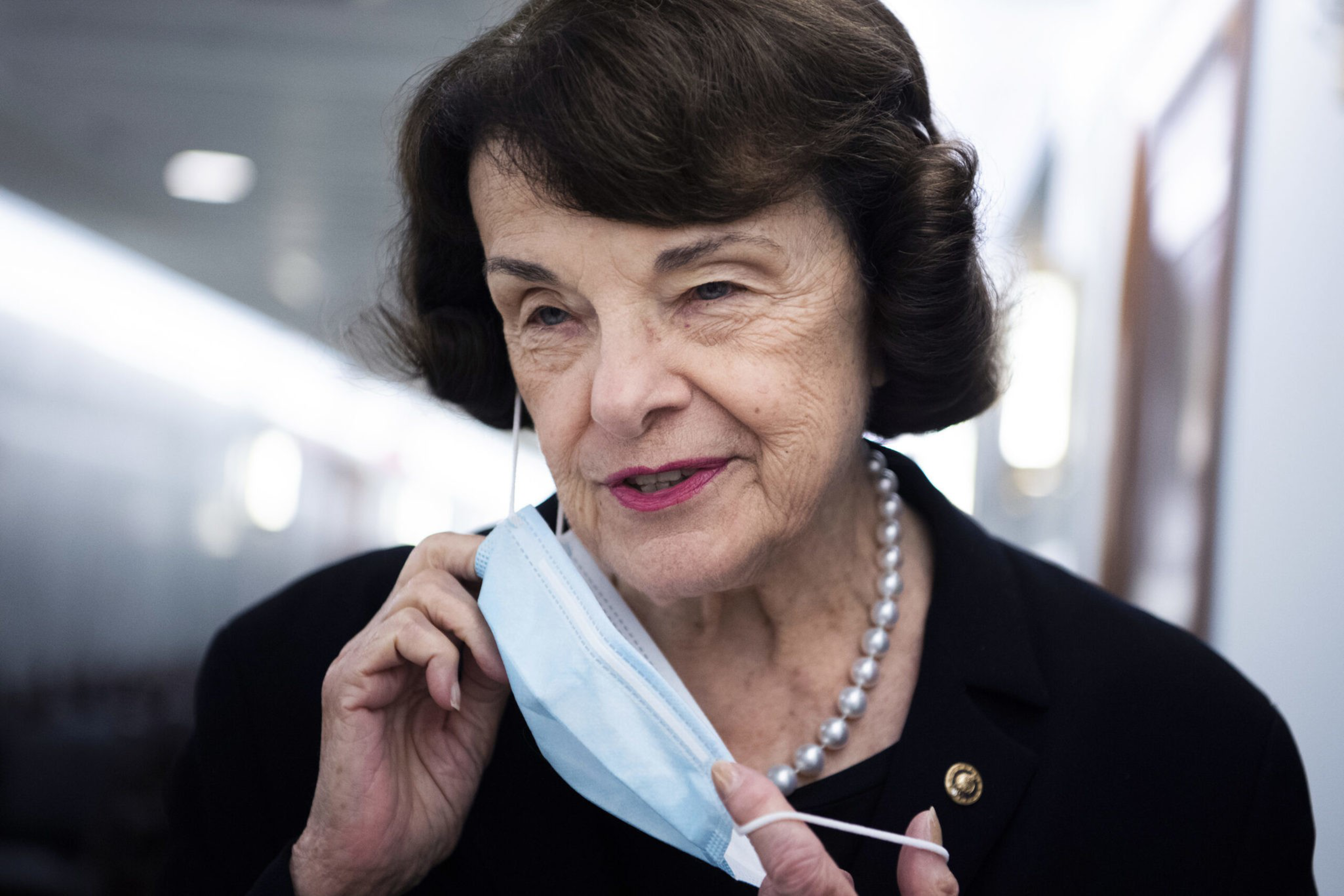 An elderly woman with short, dark hair is taking off a blue mask. She's wearing a black suit with a pearl necklace and a small pin, standing in a bright hallway.